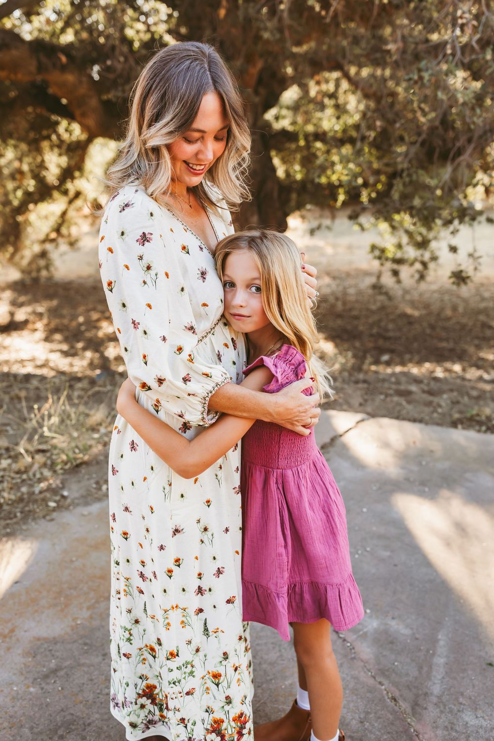A mother and daughter in coordinated floral dress and pink romper share tender moments outdoors in natural lighting.
