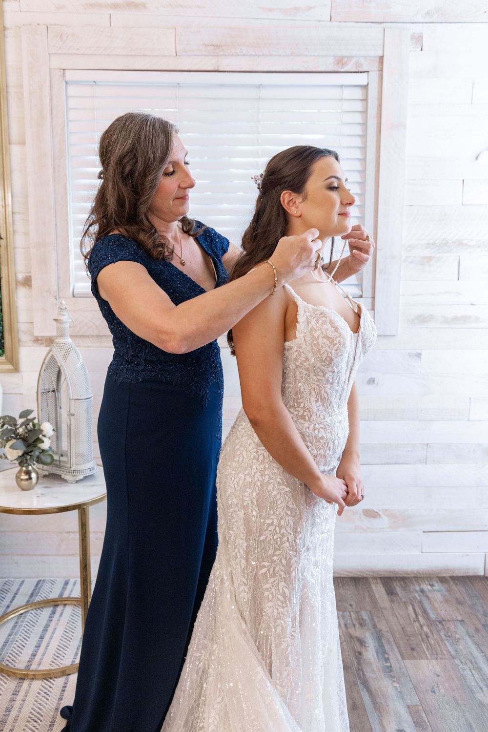 Woman in navy blue dress helps bride get ready in white lace wedding gown against rustic white wall.