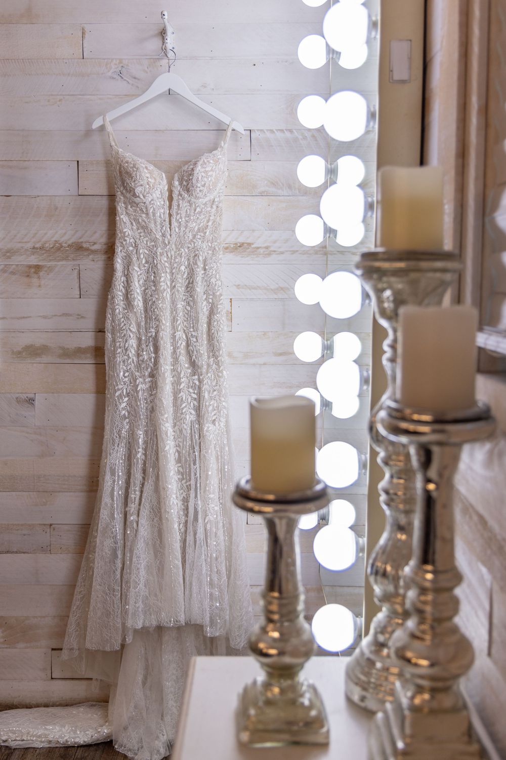 Elegant white lace wedding dress hanging in a dressing room with illuminated mirrors and decorative candle holders.