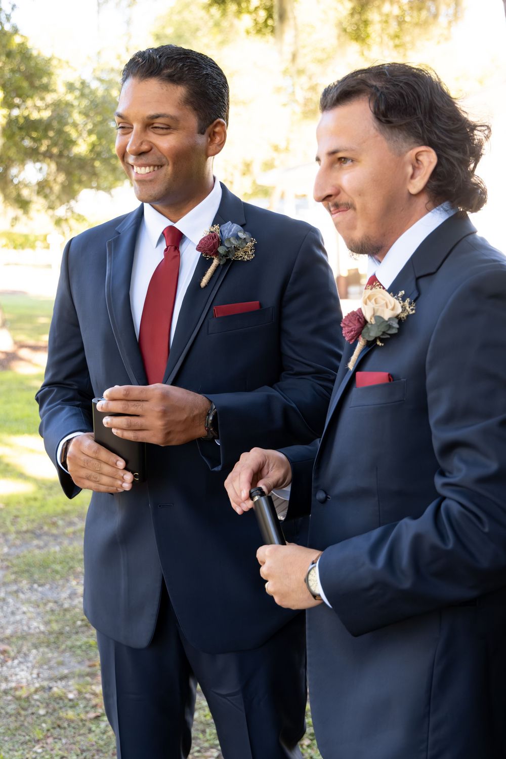Groomsmen in navy suits with red ties share laughs and drinks during outdoor wedding celebration.