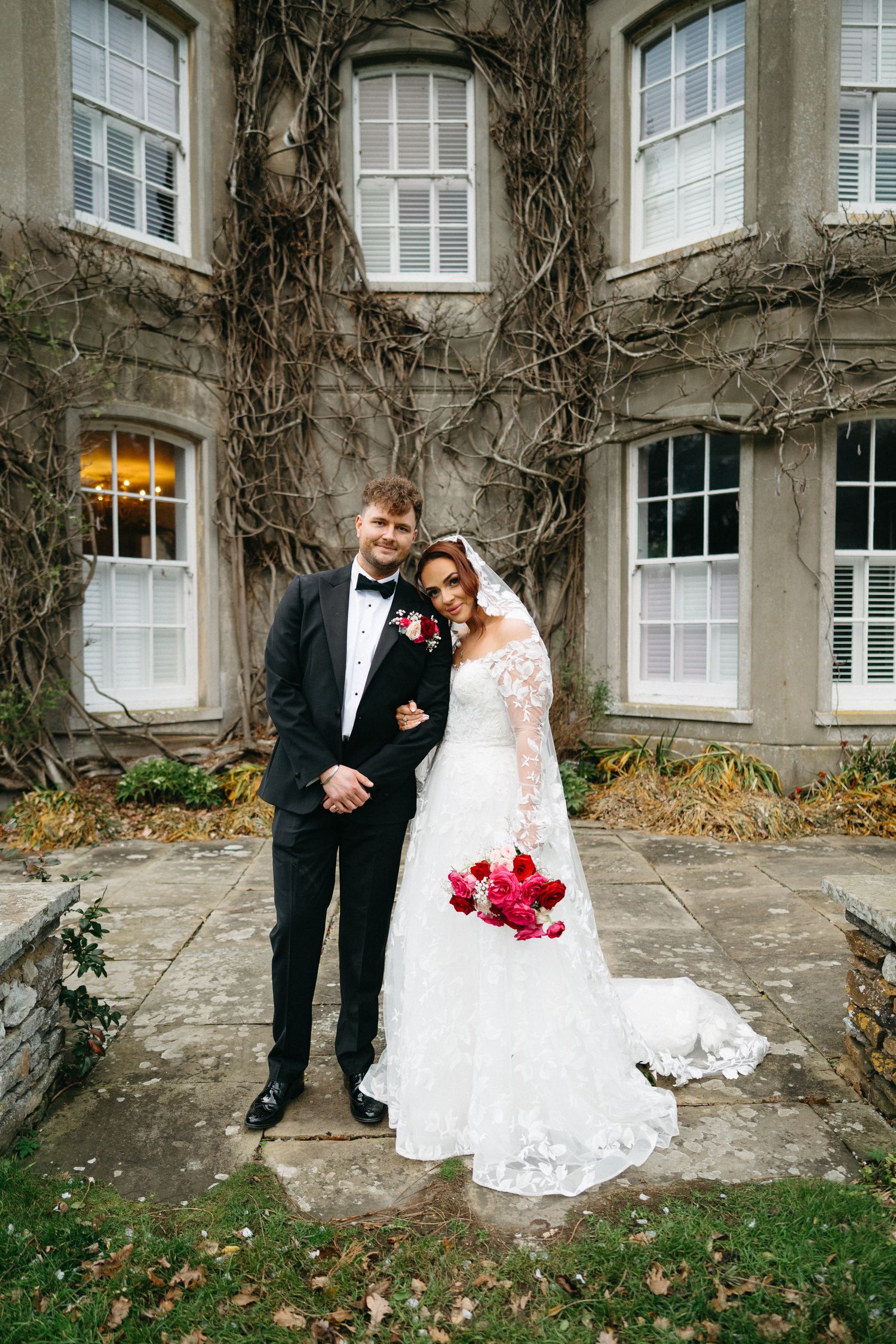 A bride in a white lace gown and groom in a black tuxedo pose together against a stone wall with climbing vines.