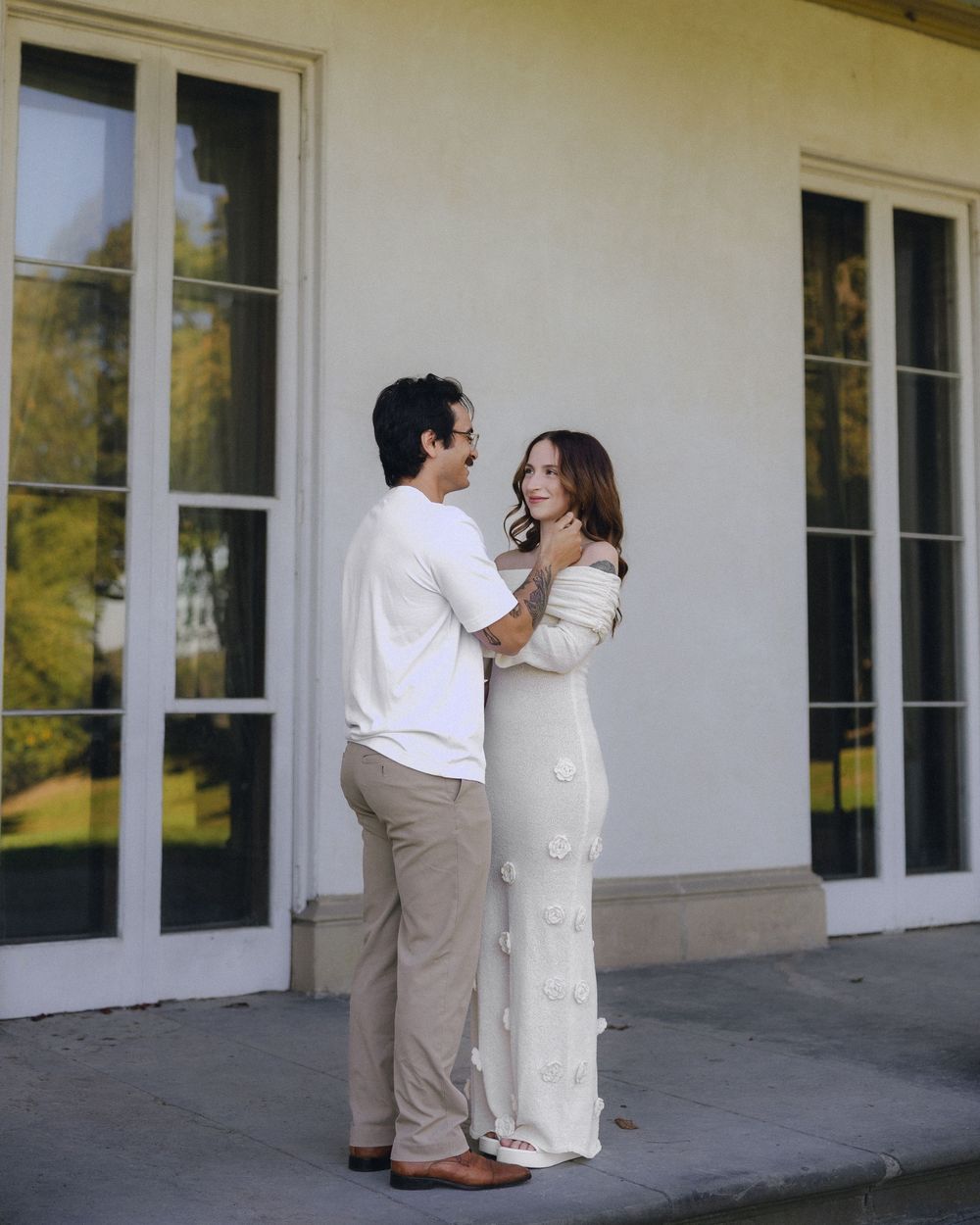 A couple in neutral tones share a romantic moment outside a white building with large windows.