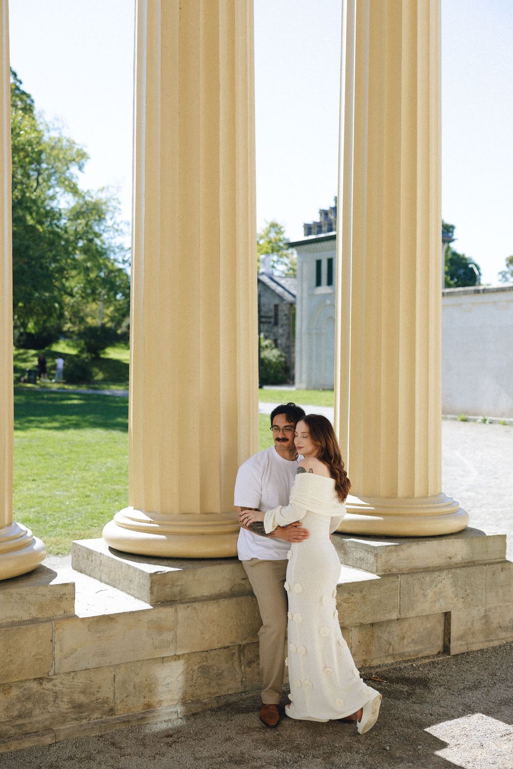 Couple in white attire embracing between tall classical columns at an elegant outdoor venue on a sunny day.