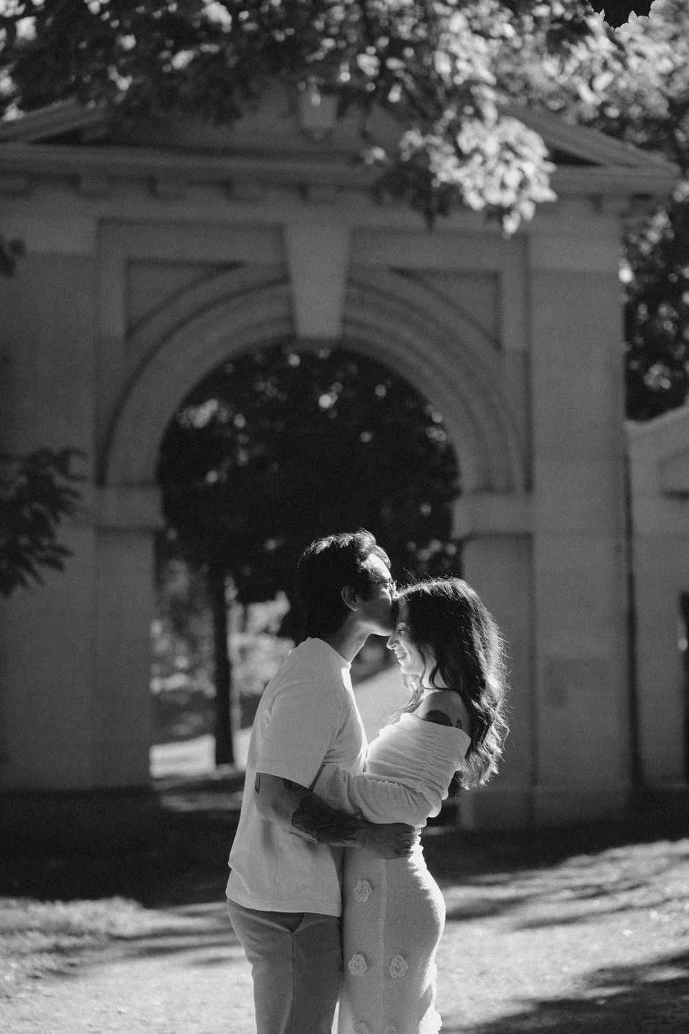 A couple embraces in a romantic outdoor setting with sunlit trees and architectural elements in the background.