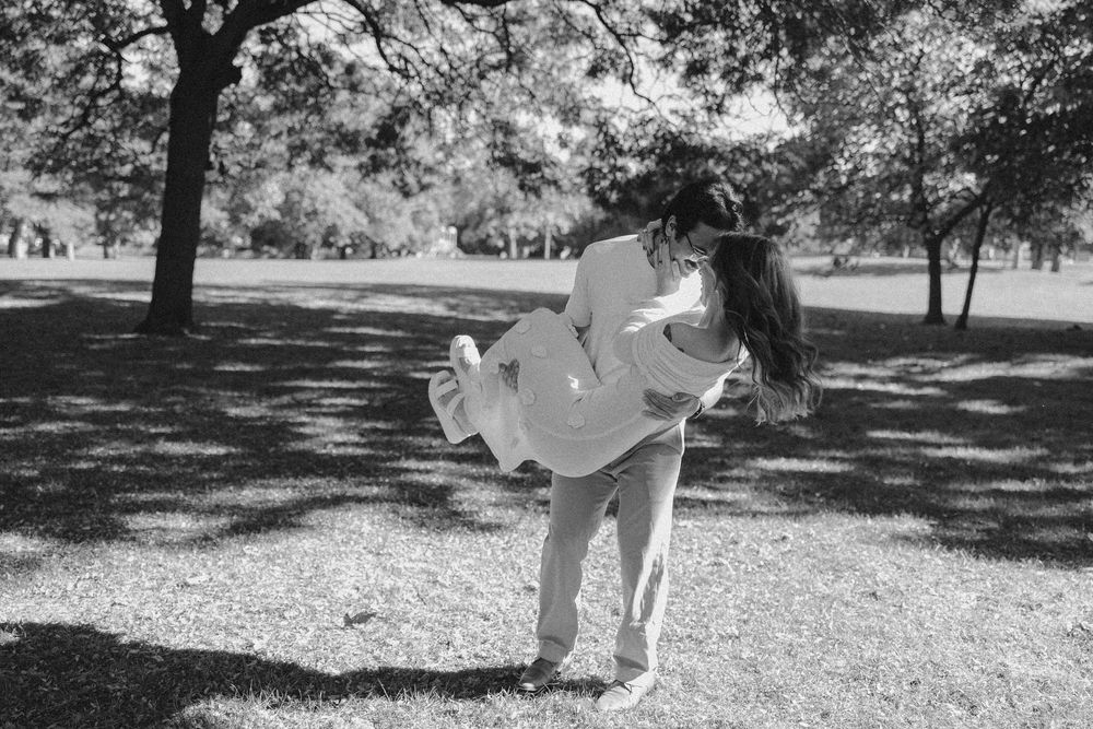 Couple sharing a romantic embrace in a sunlit park, black and white photo capturing a joyful lift and kiss.