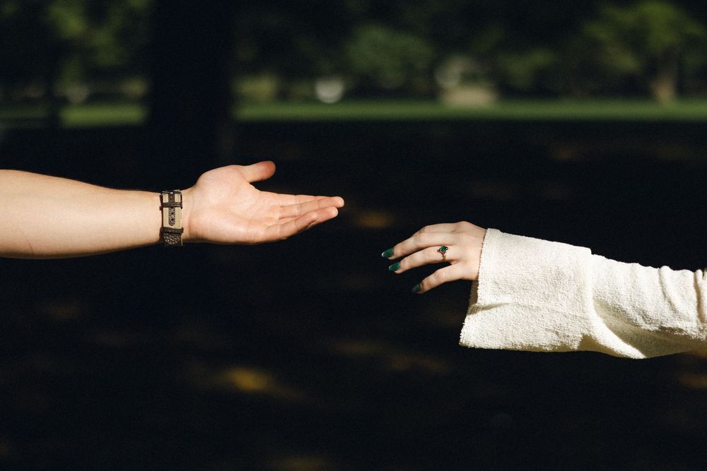 Two hands reaching toward each other outdoors, one wearing a bracelet and one wearing a ring, symbolizing connection.