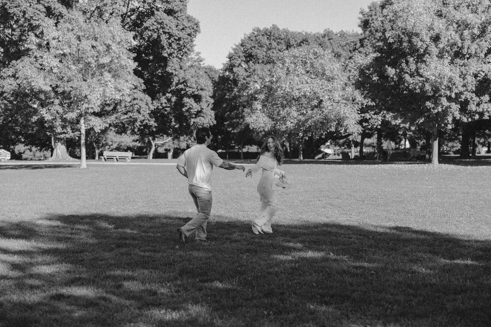 Two people holding hands and running through a lush green park on a sunny day, captured in black and white.