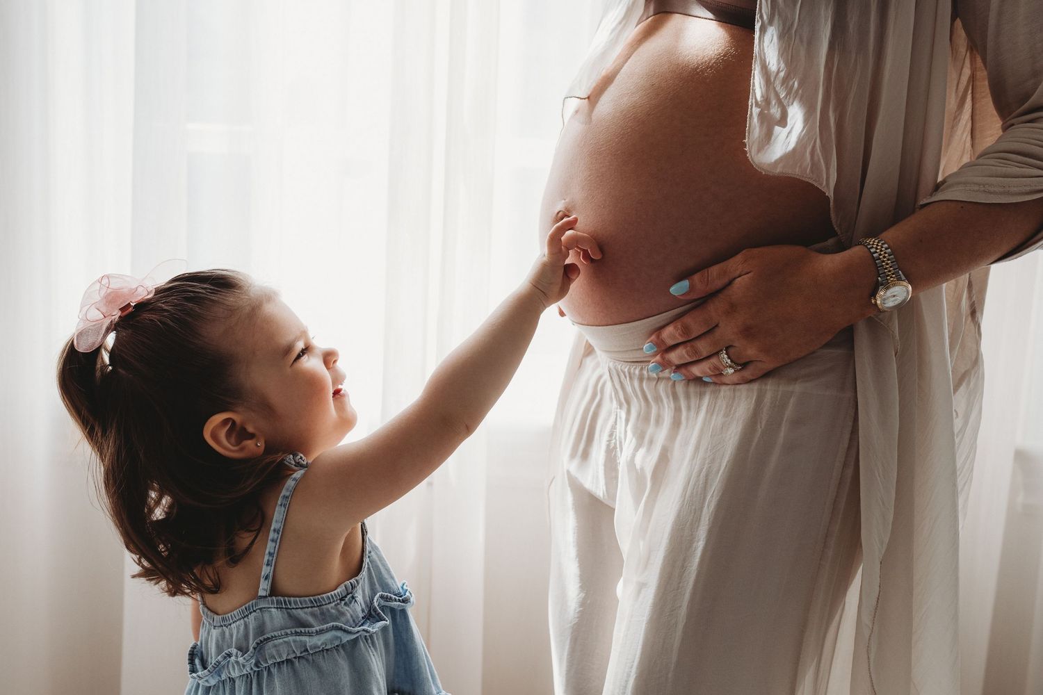 Young child in denim overalls gently touches pregnant woman's belly near a window with soft lighting.