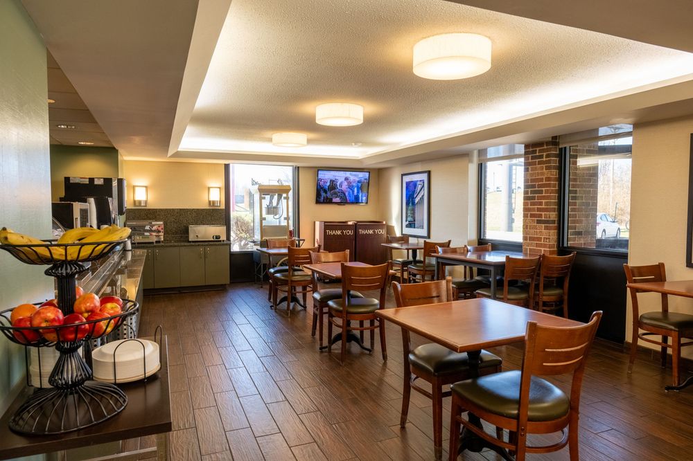 A modern hotel dining area with multiple tables and chairs illuminated by ceiling lights and natural window lighting.