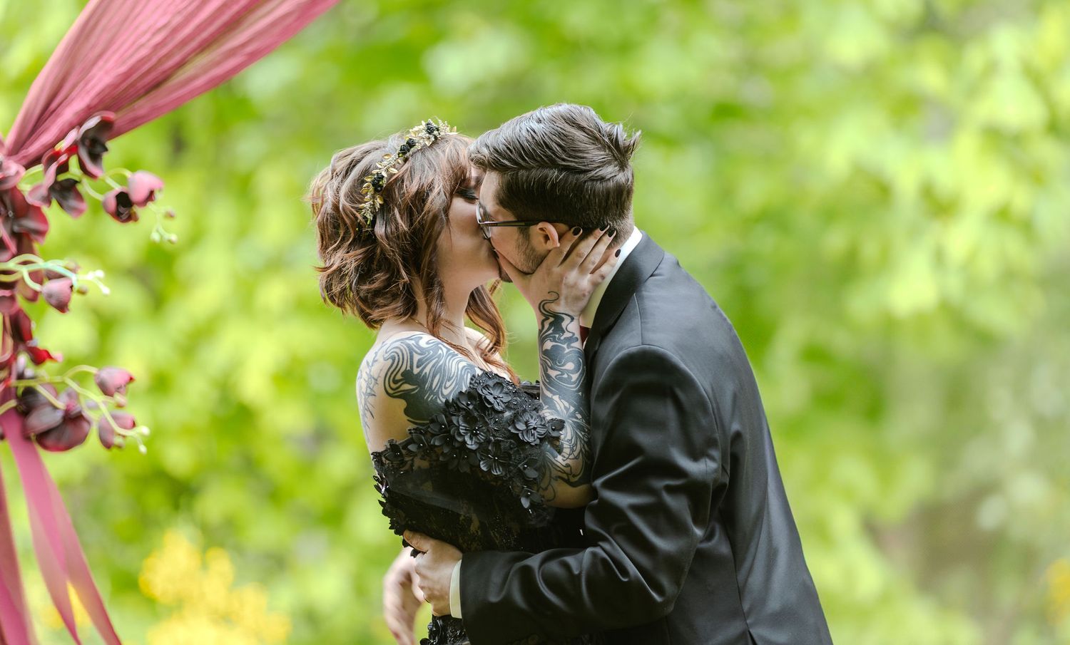 Bride and groom sharing first kiss at outdoor wedding ceremony under burgundy floral arch.