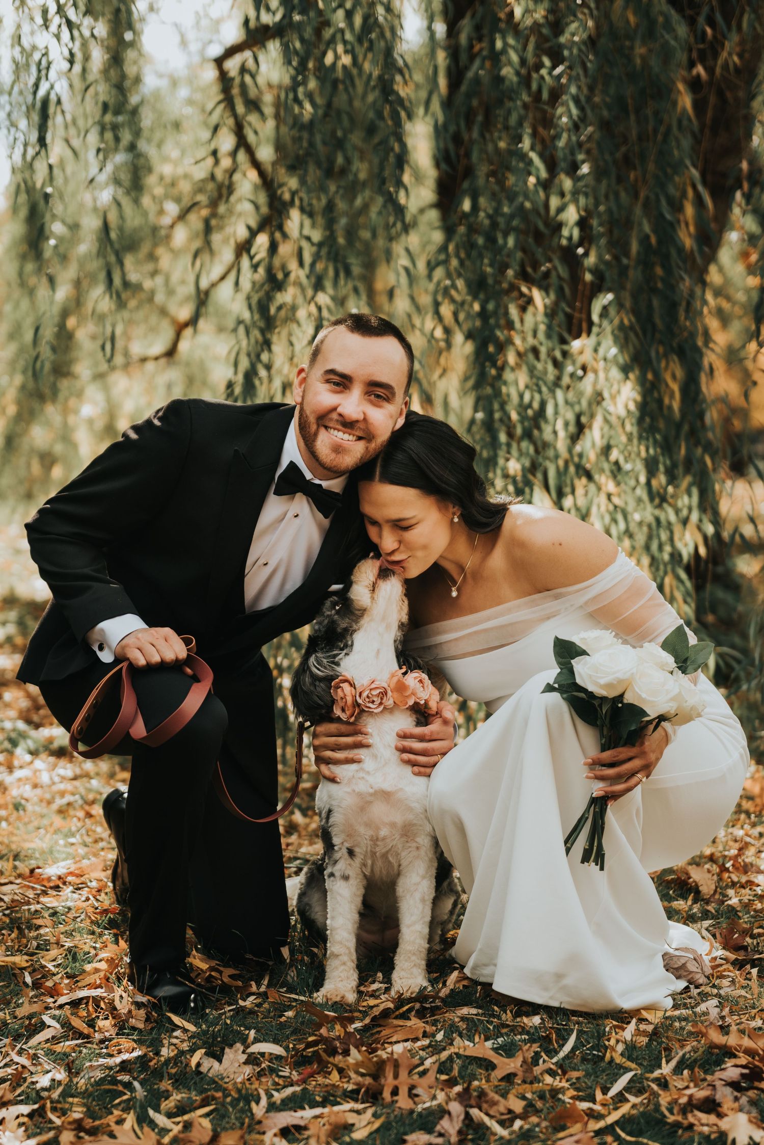 A romantic wedding couple shares playful moments among willow trees and fallen autumn leaves.