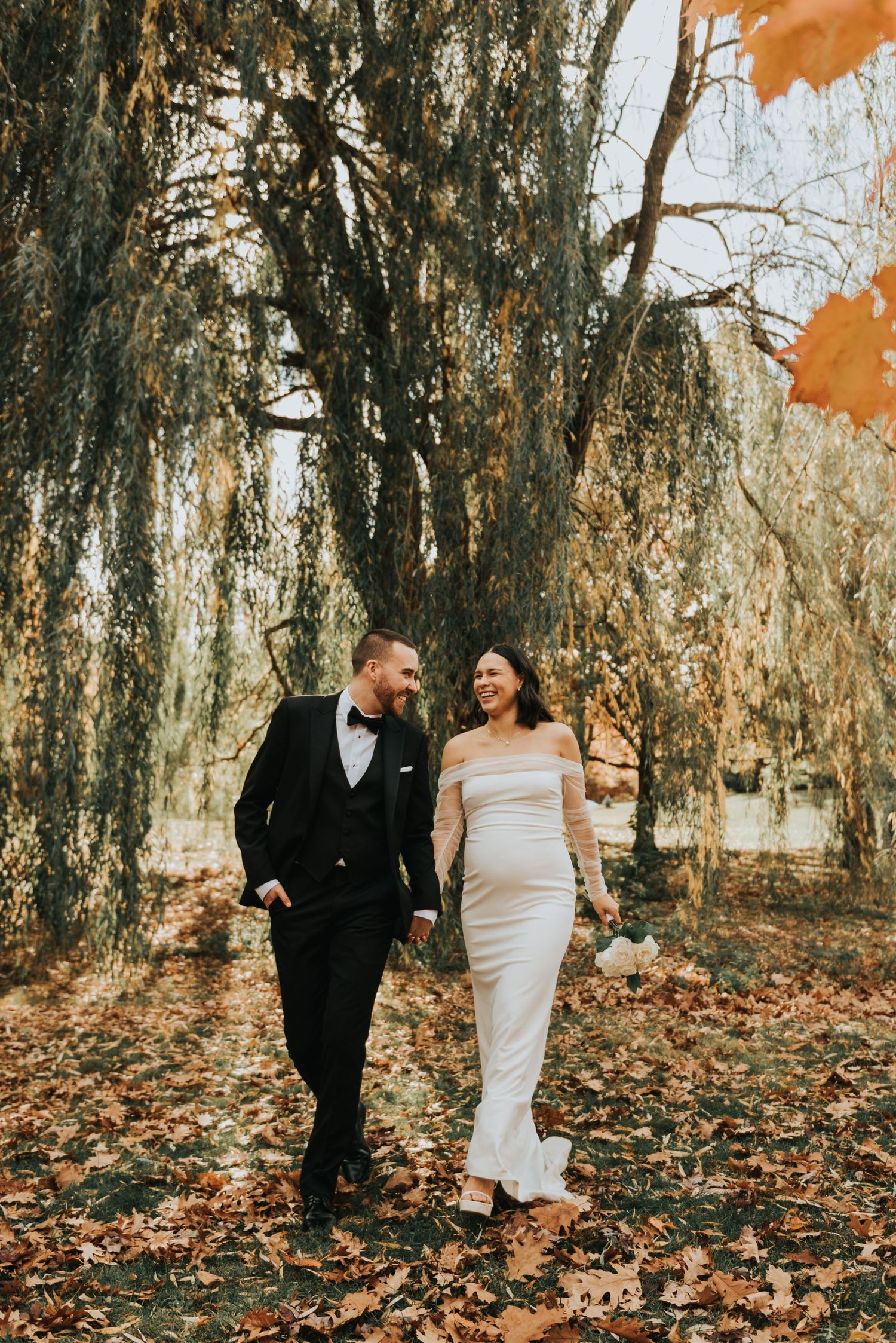 A romantic autumn wedding photo of a couple walking through willow trees with orange leaves falling.