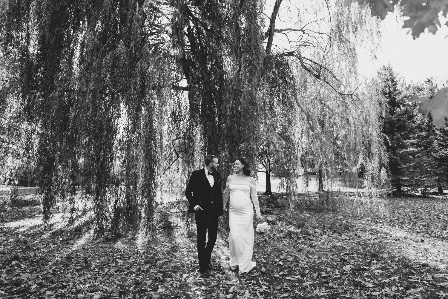 A couple in wedding attire stands beneath a dramatic weeping willow tree in a black and white photograph.