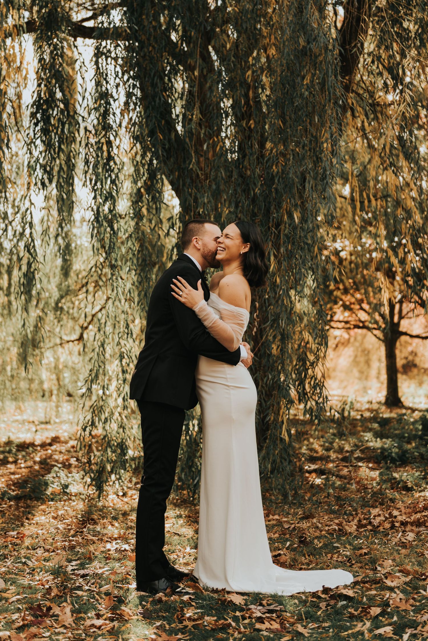A romantic wedding couple embraces under weeping willow trees in an autumn setting.