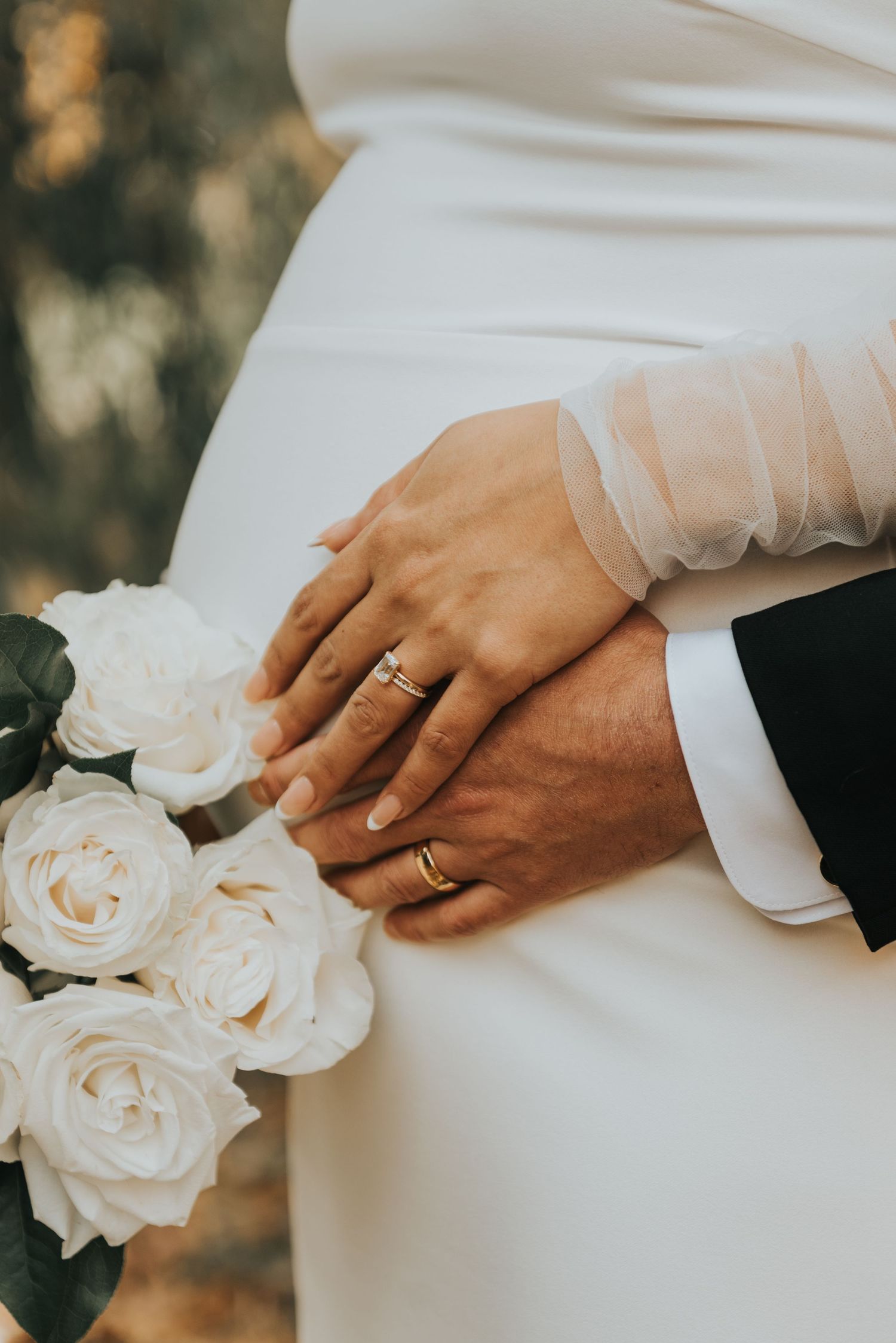 Close-up of newlywed couple's hands with wedding rings and white roses against bride's white dress.