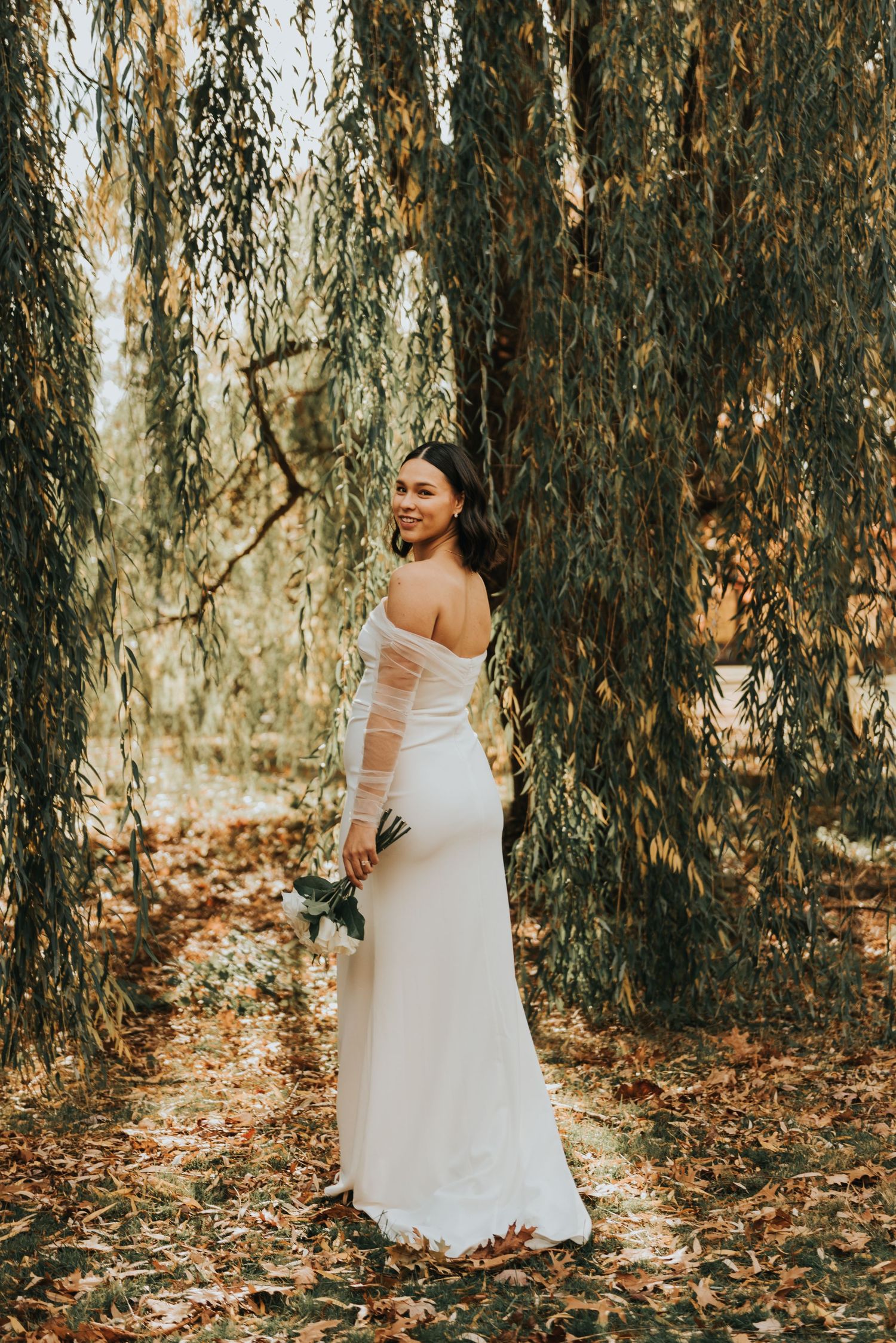 Woman in flowing white dress poses gracefully among autumn willow trees with fallen leaves.