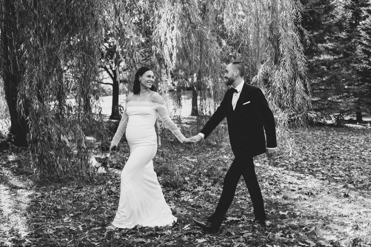 A couple in wedding attire walks hand in hand beneath a dramatic weeping willow tree in black and white.