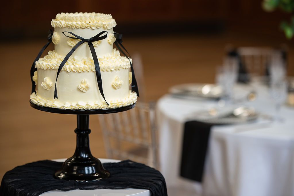 White tiered wedding cake with black ribbon sits on a stand, made with sustainable ingredients