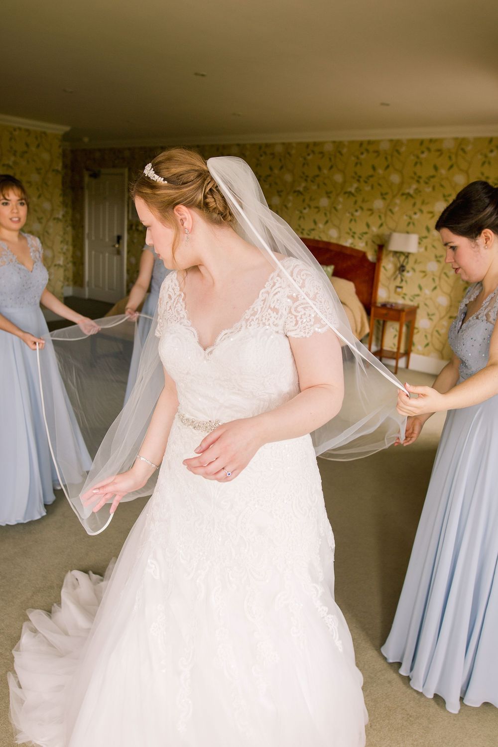 Wedding preparation scene showing light blue bridesmaid dresses and a white wedding gown in a floral wallpapered room.