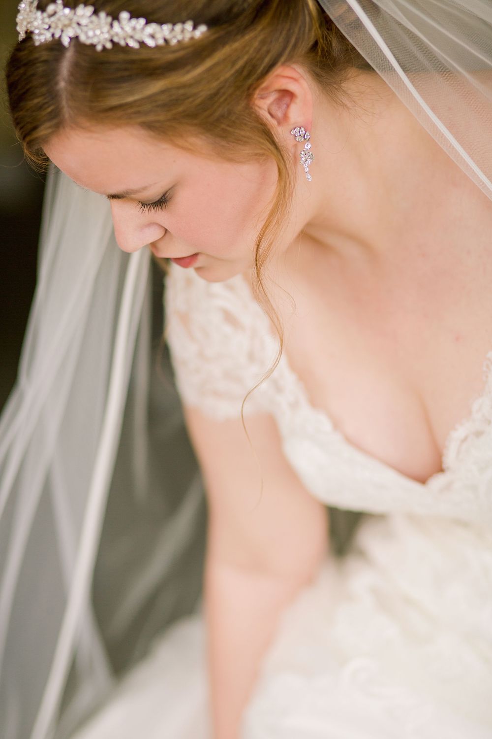 Bride in white lace wedding dress with delicate crystal earrings looks down pensively in romantic portrait series.