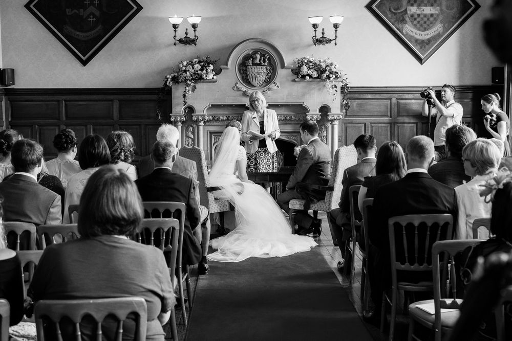 A black and white photograph of a wedding ceremony taking place in an intimate indoor venue with ornate decor.