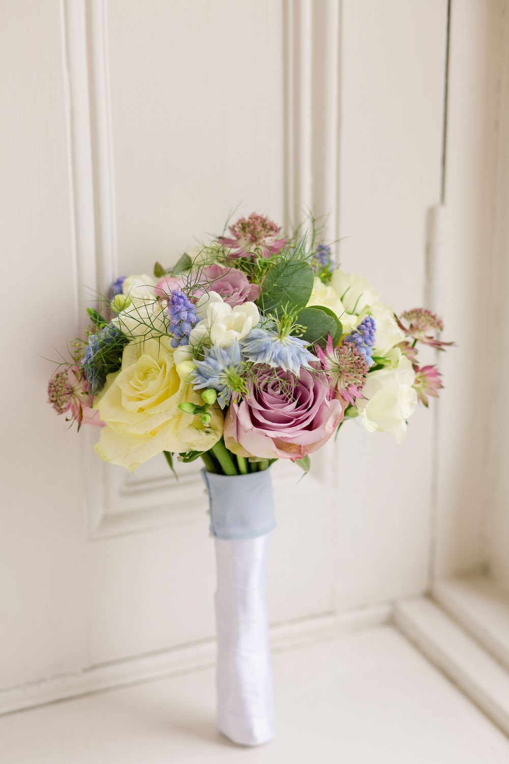 A series of delicate floral arrangements in white vases featuring yellow roses, purple blooms, and greenery against a white wall.