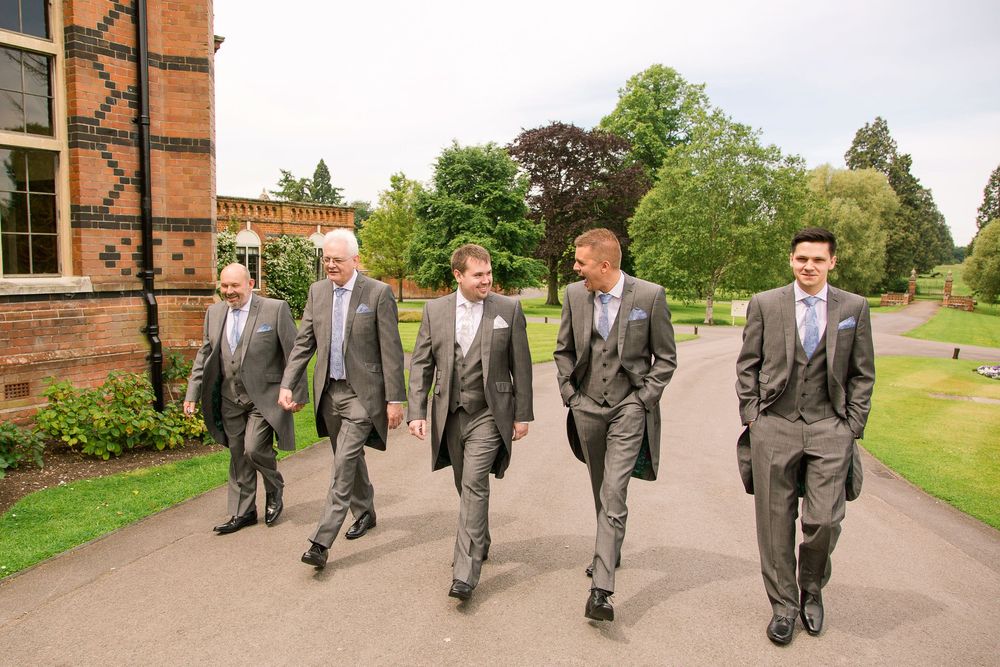 Group walking along a paved path beside red brick buildings wearing matching grey suits.