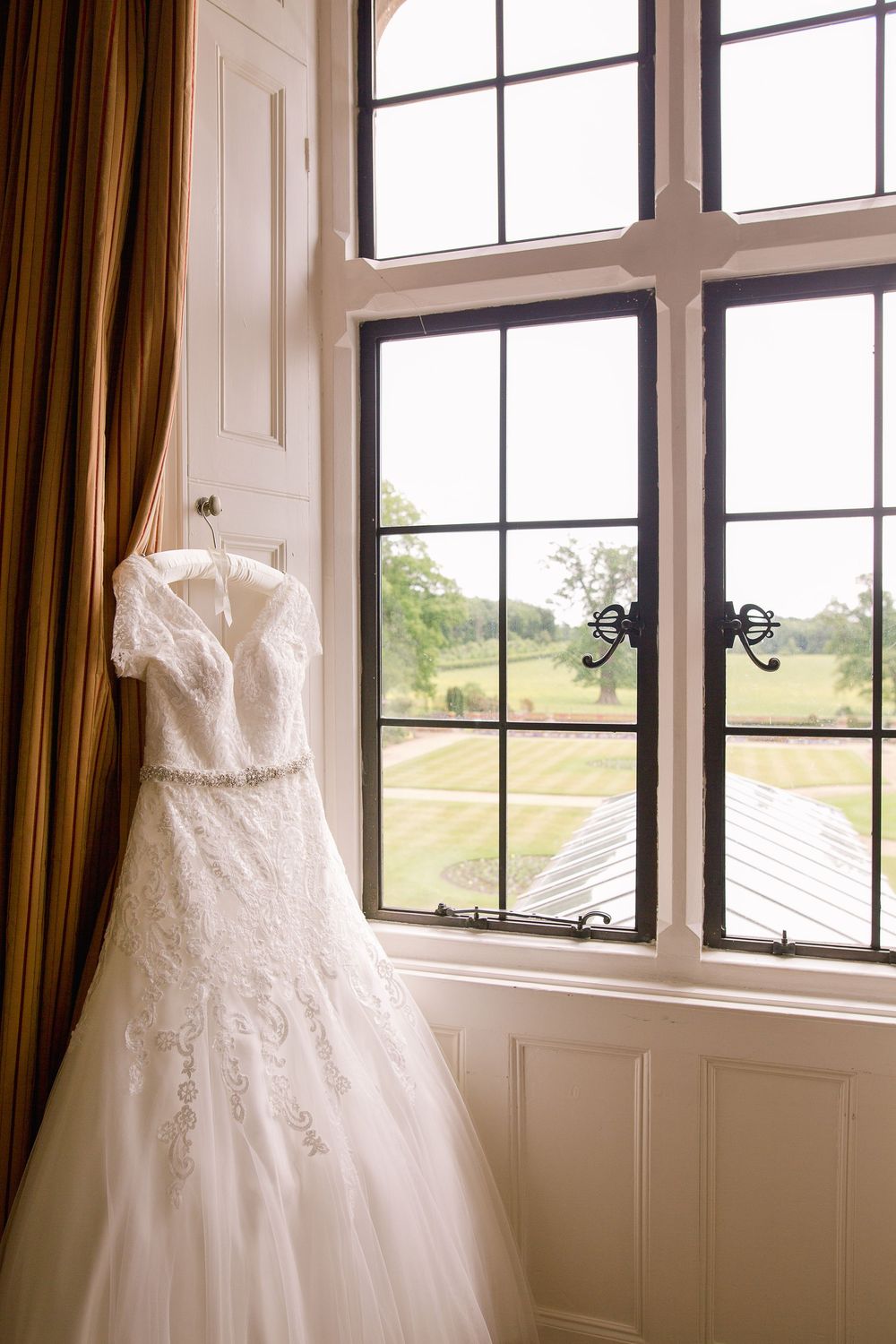 A white lace wedding dress hangs by a large window with a scenic countryside view and brown curtains.