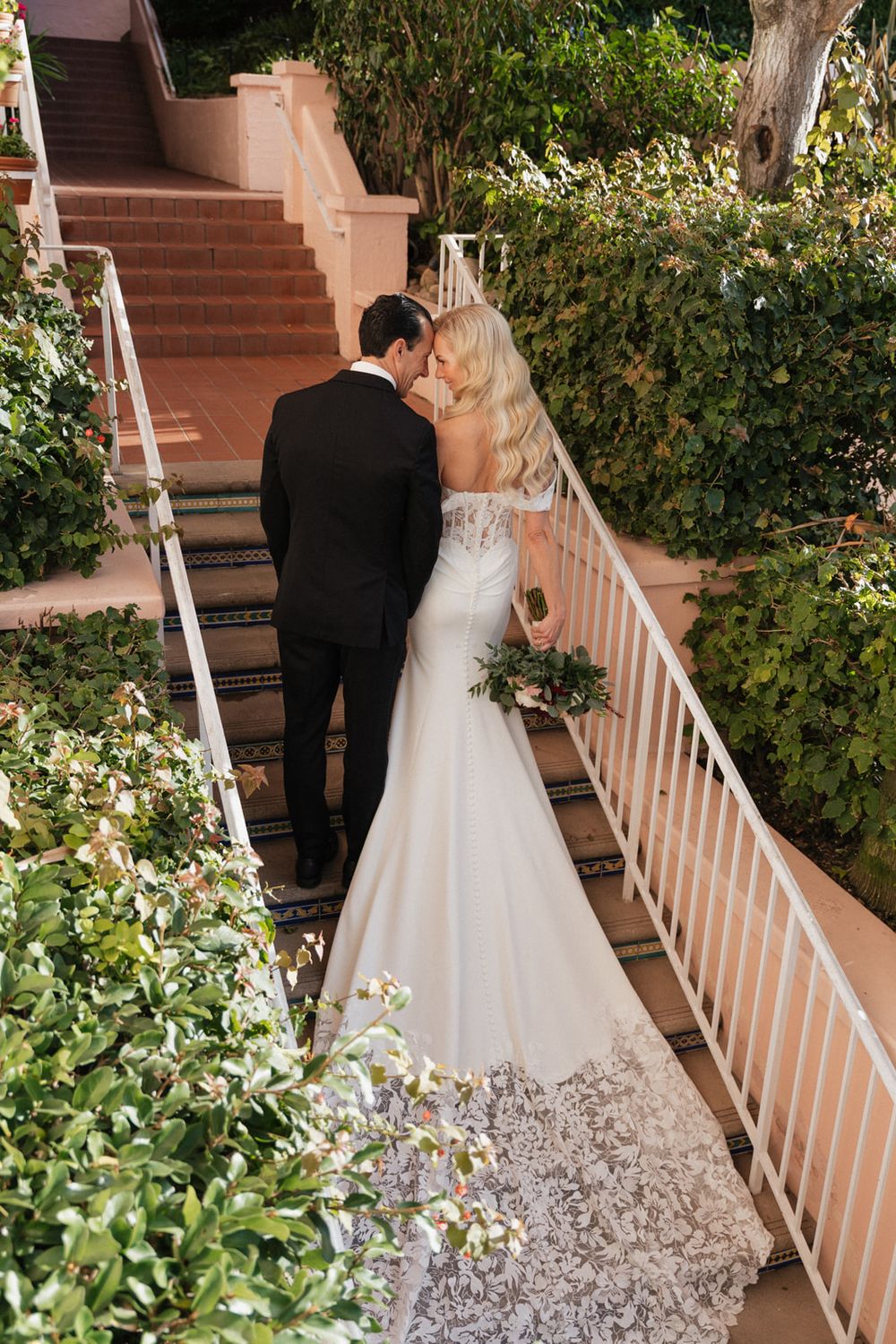 A couple in wedding attire ascends outdoor stairs surrounded by lush greenery and white railings.