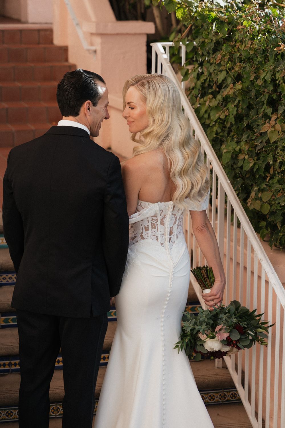 A romantic wedding couple shares an intimate moment on a staircase with brick walls and greenery in the background.
