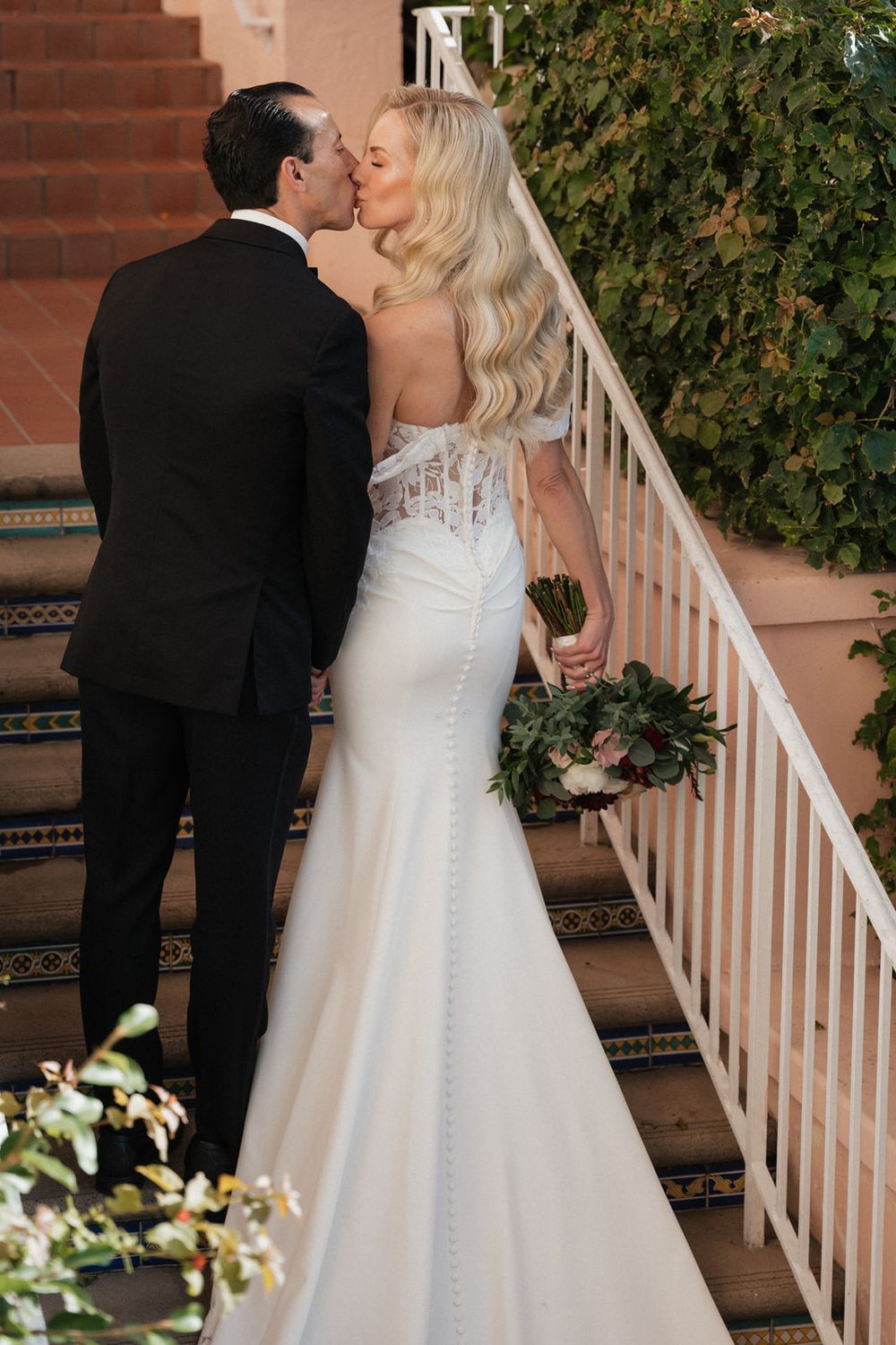 A couple shares an intimate moment on stairs, with the bride wearing a fitted white gown and holding a bouquet.