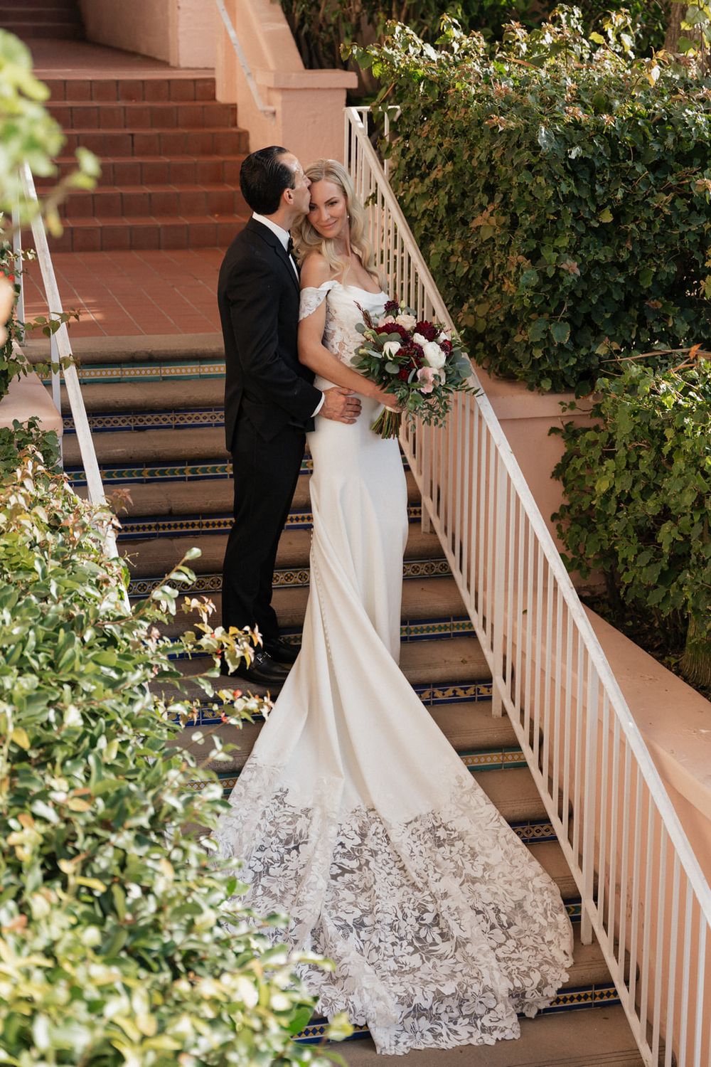 Newlywed couple shares romantic moment on brick steps with elegant white wedding gown featuring lace train detail.