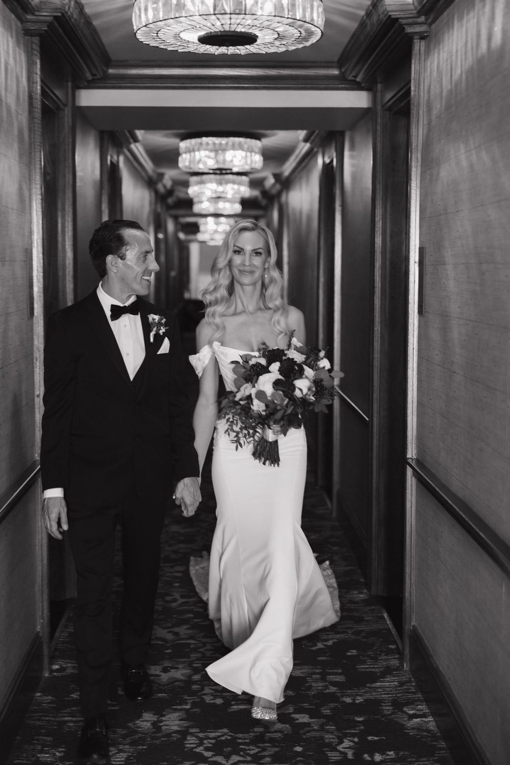 A bride and groom walk through an elegant hotel hallway with crystal chandeliers and patterned carpet.