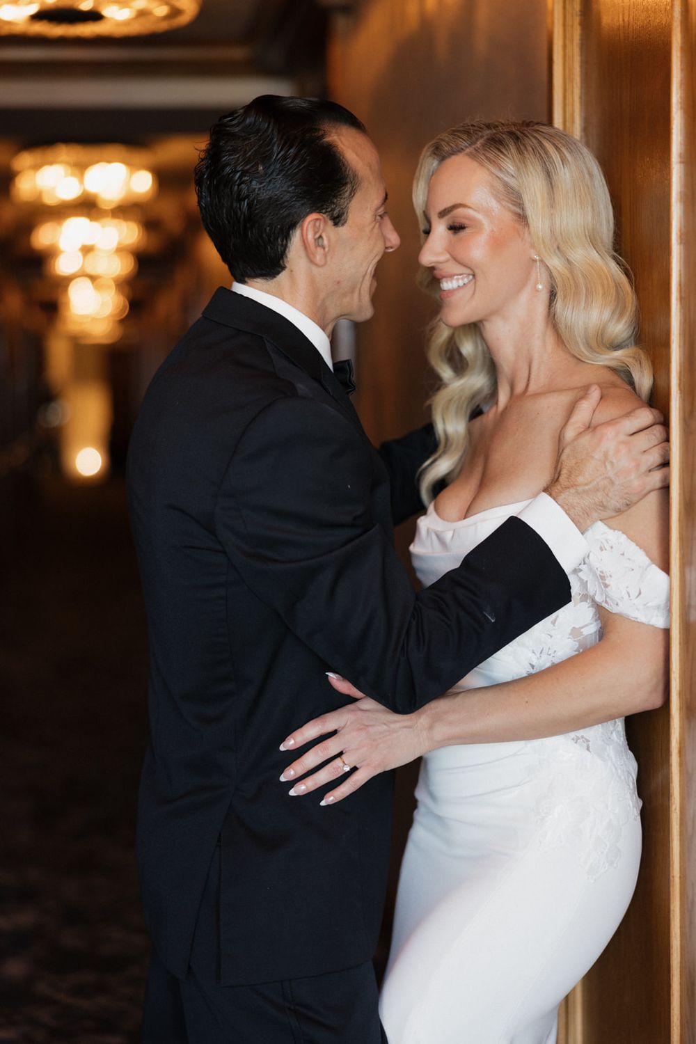 A couple in formal wedding attire share an intimate moment in an elegant indoor venue with chandelier lighting.