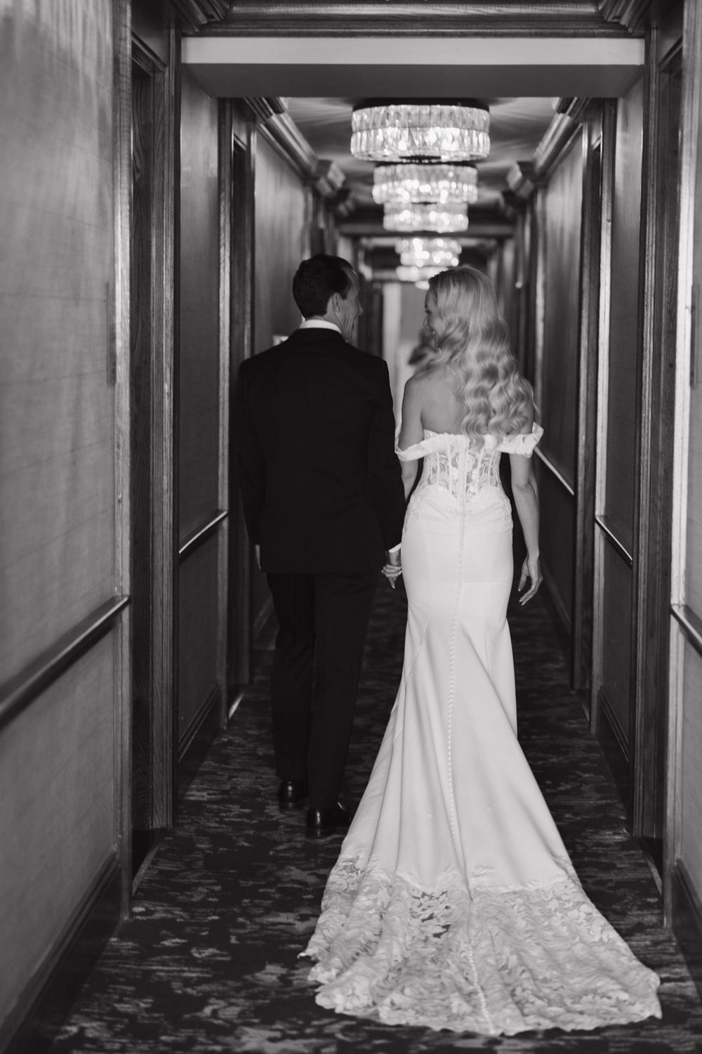 Black and white photo of a couple walking down an elegant hotel hallway with crystal chandeliers overhead.