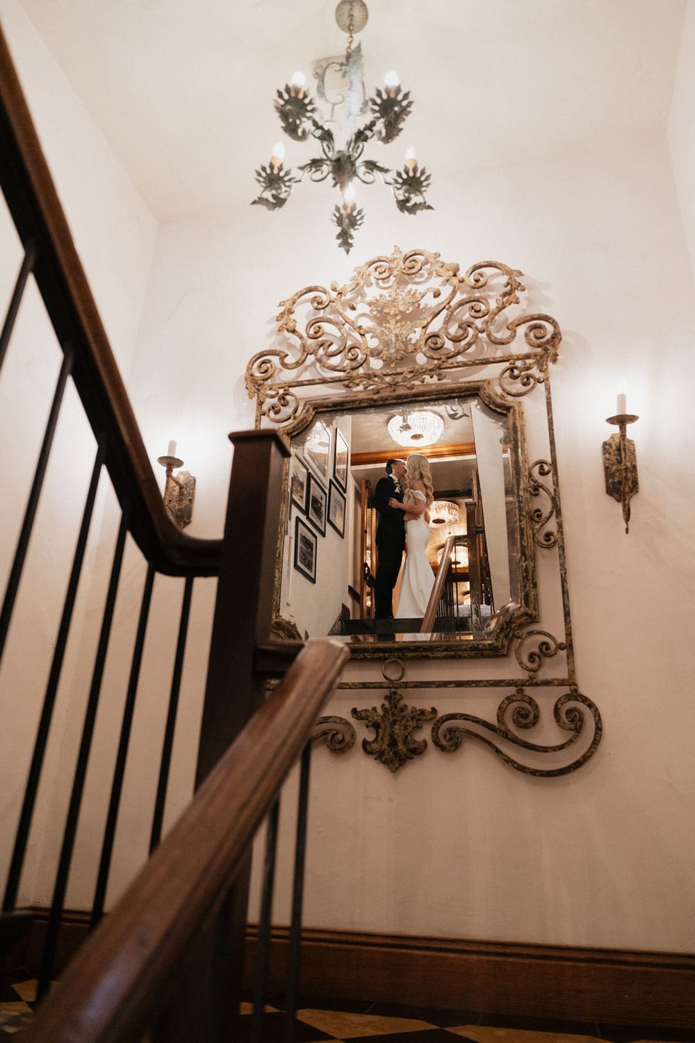 Ornate vintage mirror with decorative metalwork crown mounted on white wall beside dark wooden staircase railing.