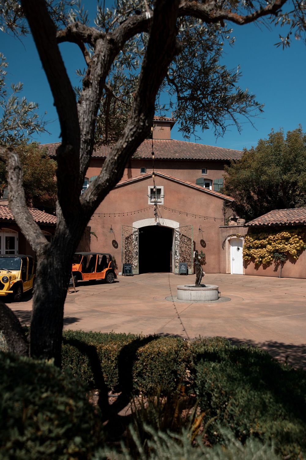 Rustic terracotta buildings with stone fountain in courtyard surrounded by mature trees and landscaping at historic estate.