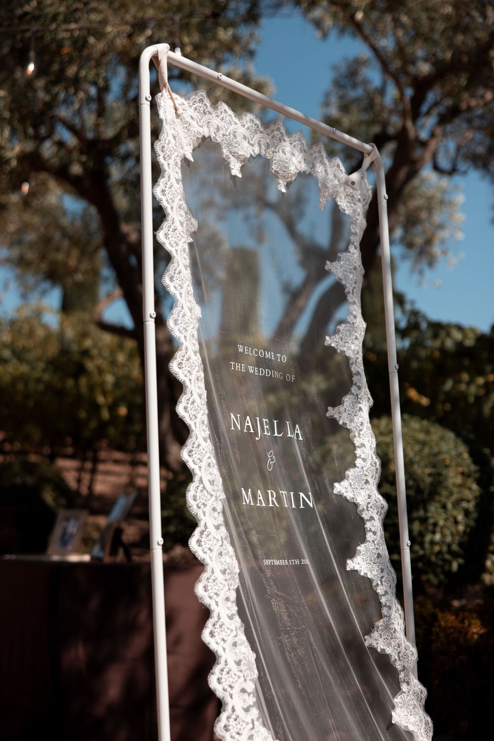 A transparent acrylic wedding sign with delicate white lace details is displayed outdoors against a natural backdrop.