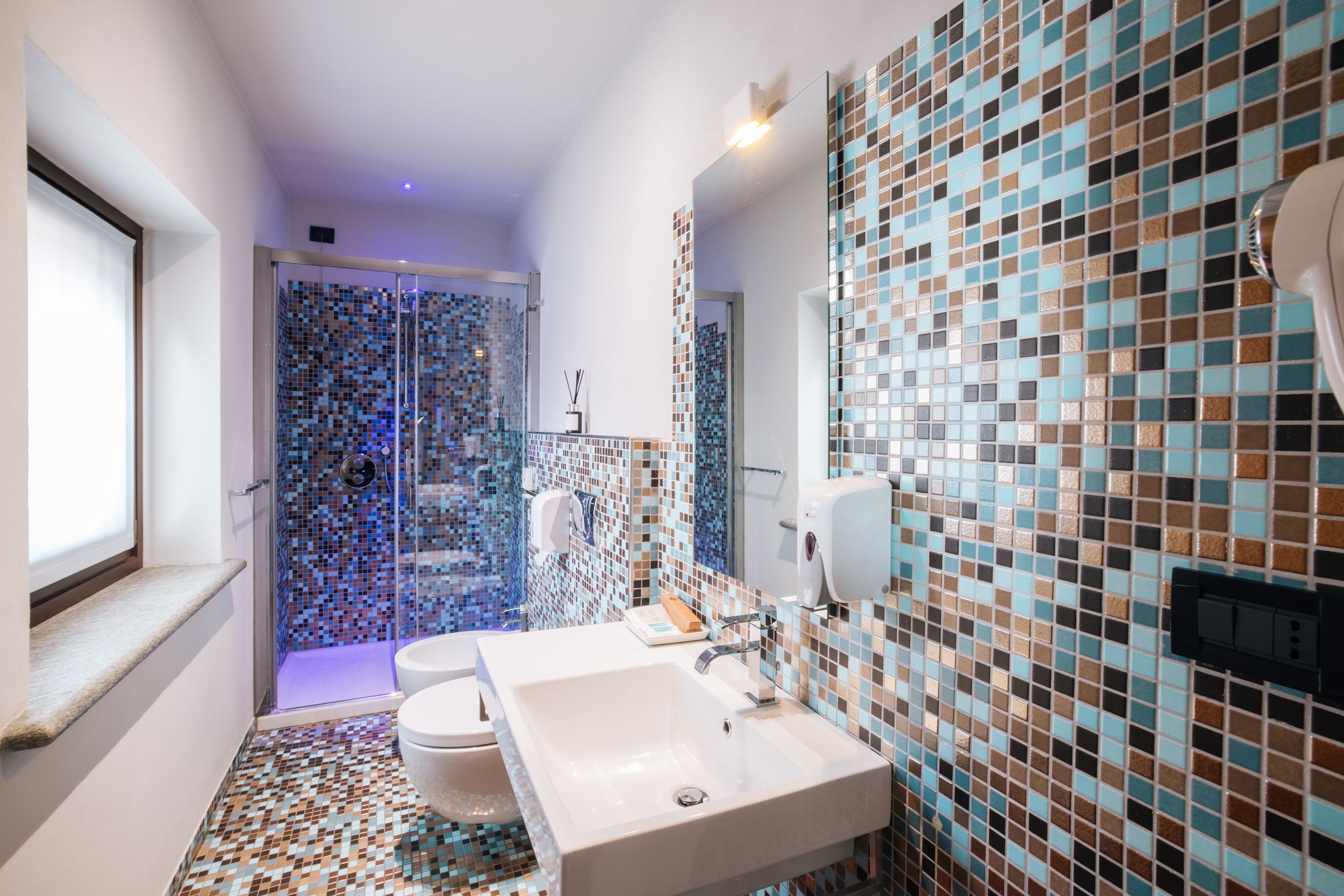 Modern bathroom interior with mosaic tile walls in blue and brown tones featuring contemporary fixtures and lighting.