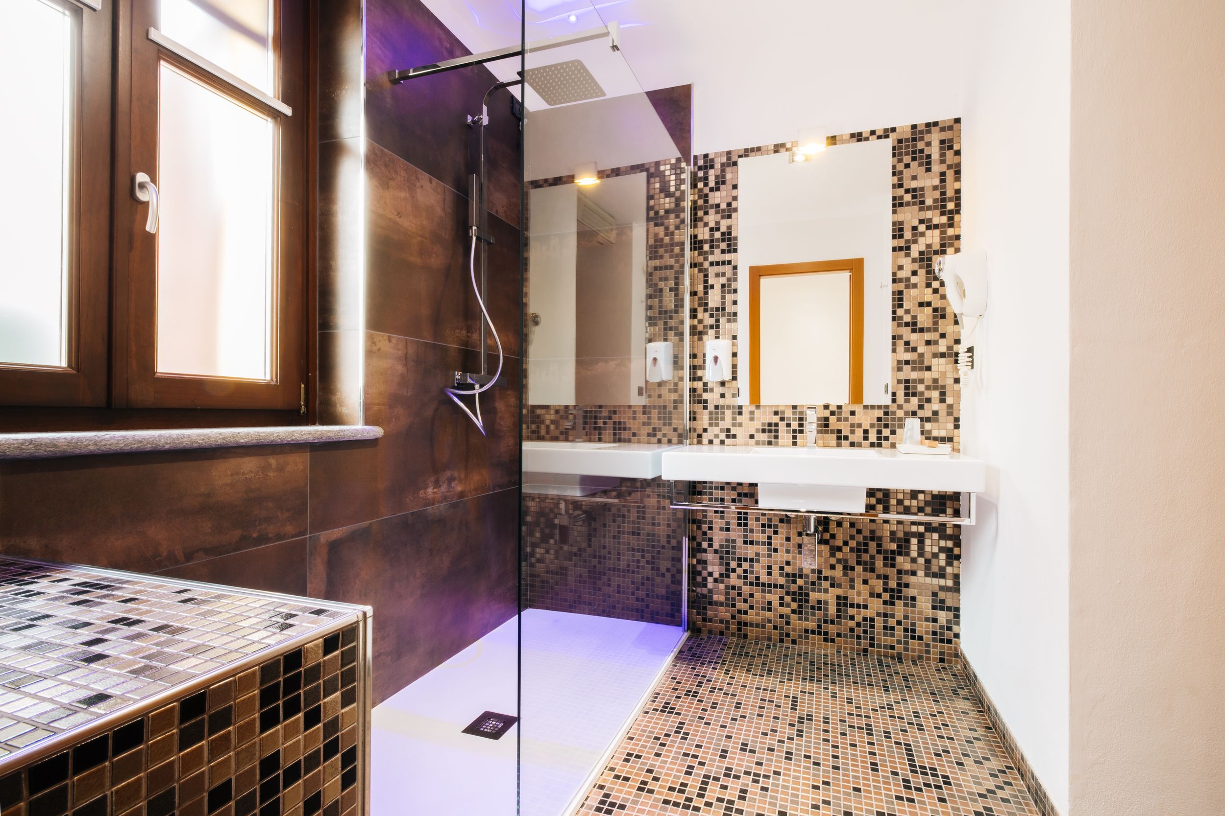 Modern bathroom interior with mosaic tile walls, wooden accents, and natural lighting from window.