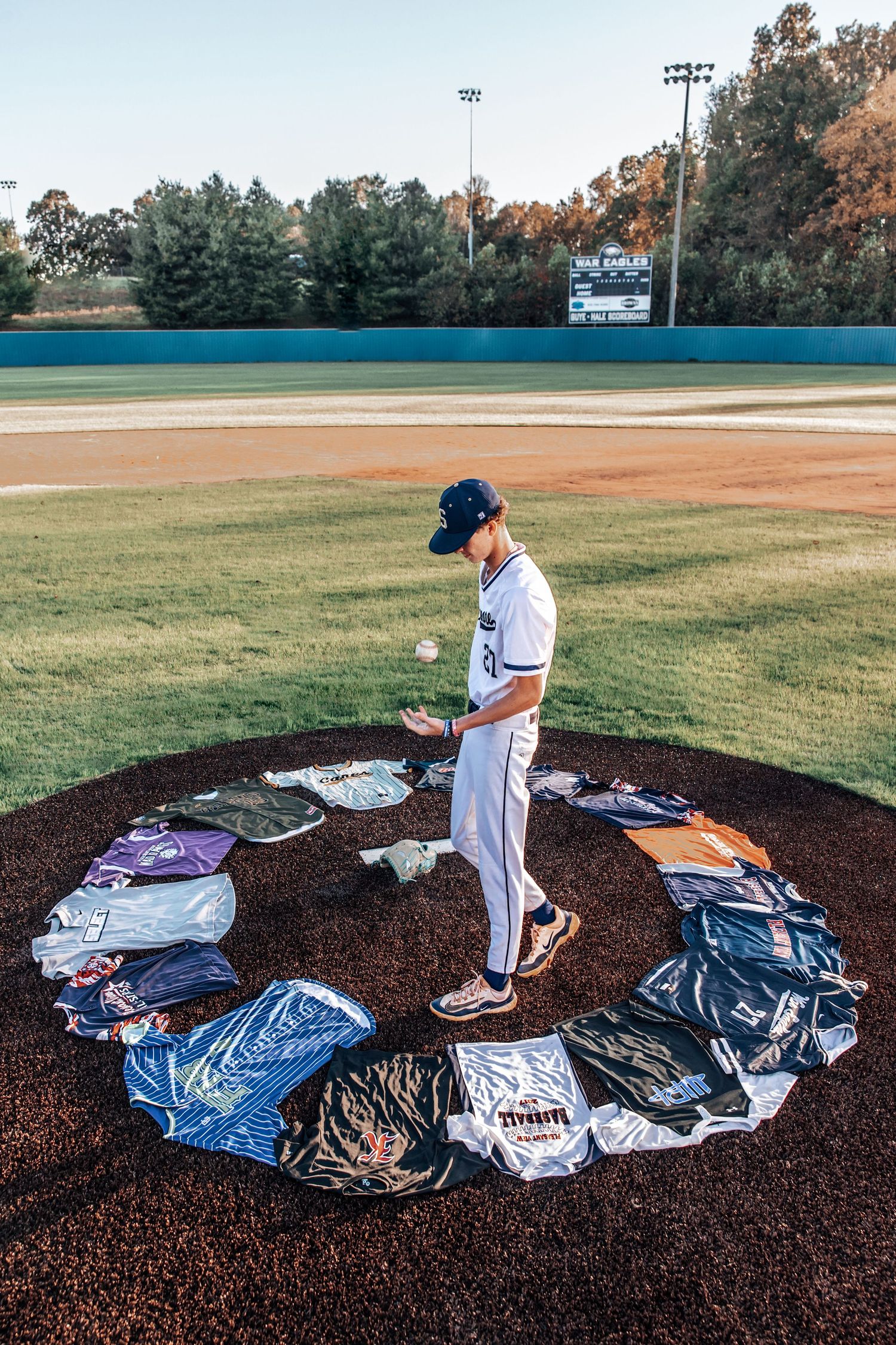 Baseball player arranges jerseys in a progressive sequence on a dirt mound at a baseball field during sunset.