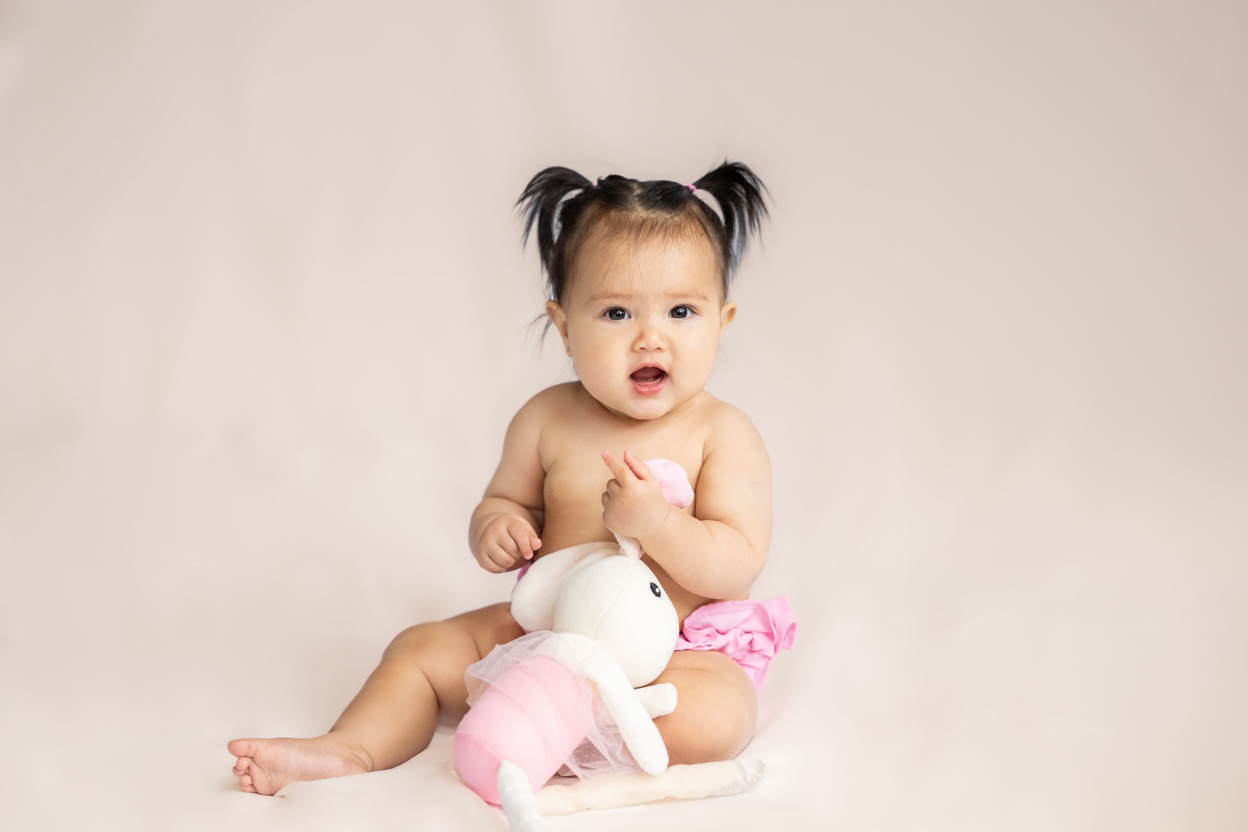Baby sitting on white backdrop with pink stuffed animal wearing hair bows in a studio in Saratoga Springs.