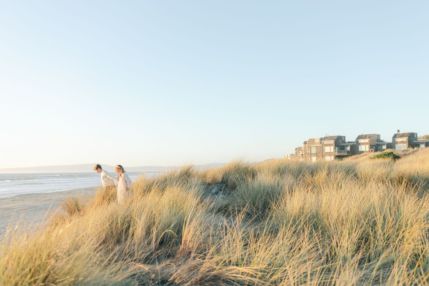 Golden beach grass in the wind along a coastal dune with beachfront homes visible in the distance beach photo session with Chelsee Rawe