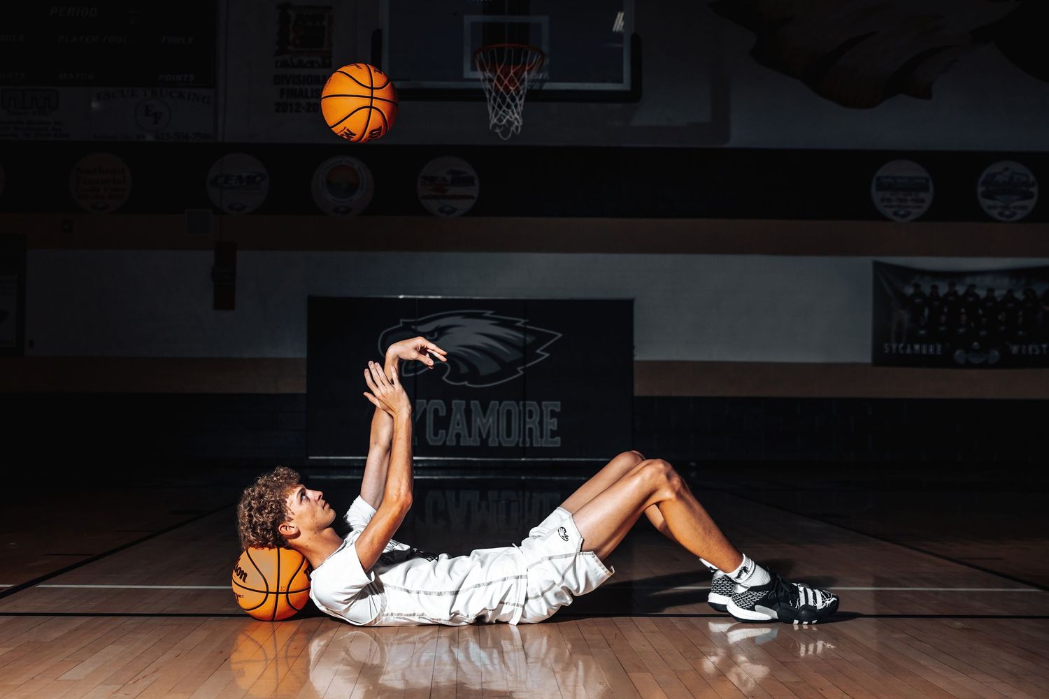 Basketball player lying on wooden court floor practicing shooting form with ball in dimly lit gymnasium.