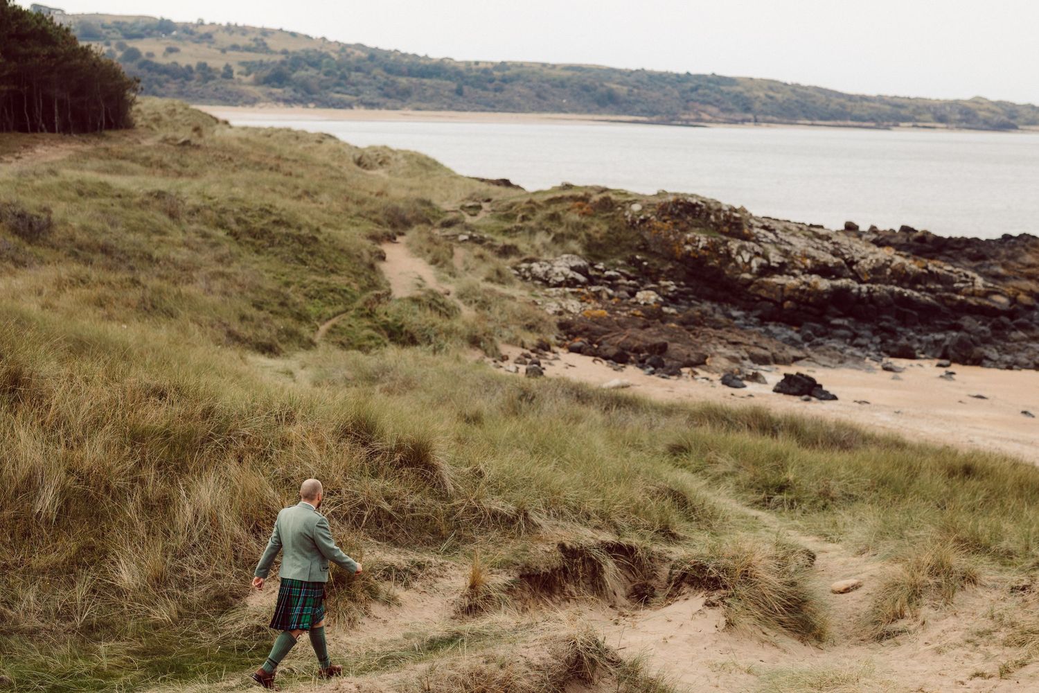 A person walks along a grassy coastal path overlooking a rocky beach and calm bay in Scotland.