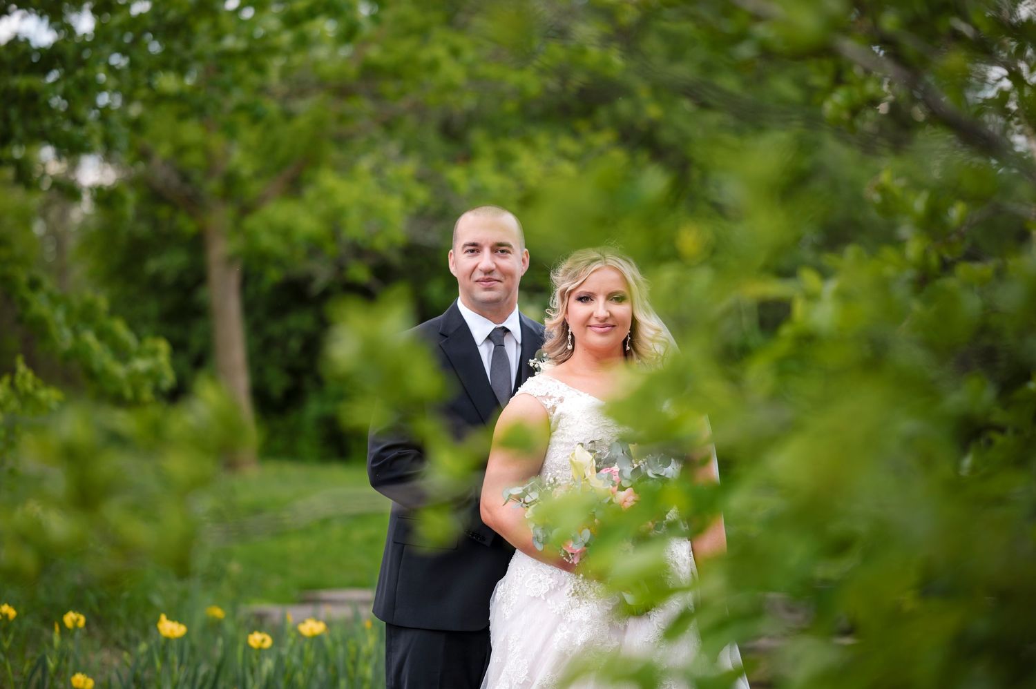 A couple in wedding attire poses for photos in a lush garden setting with vibrant greenery and flowers.