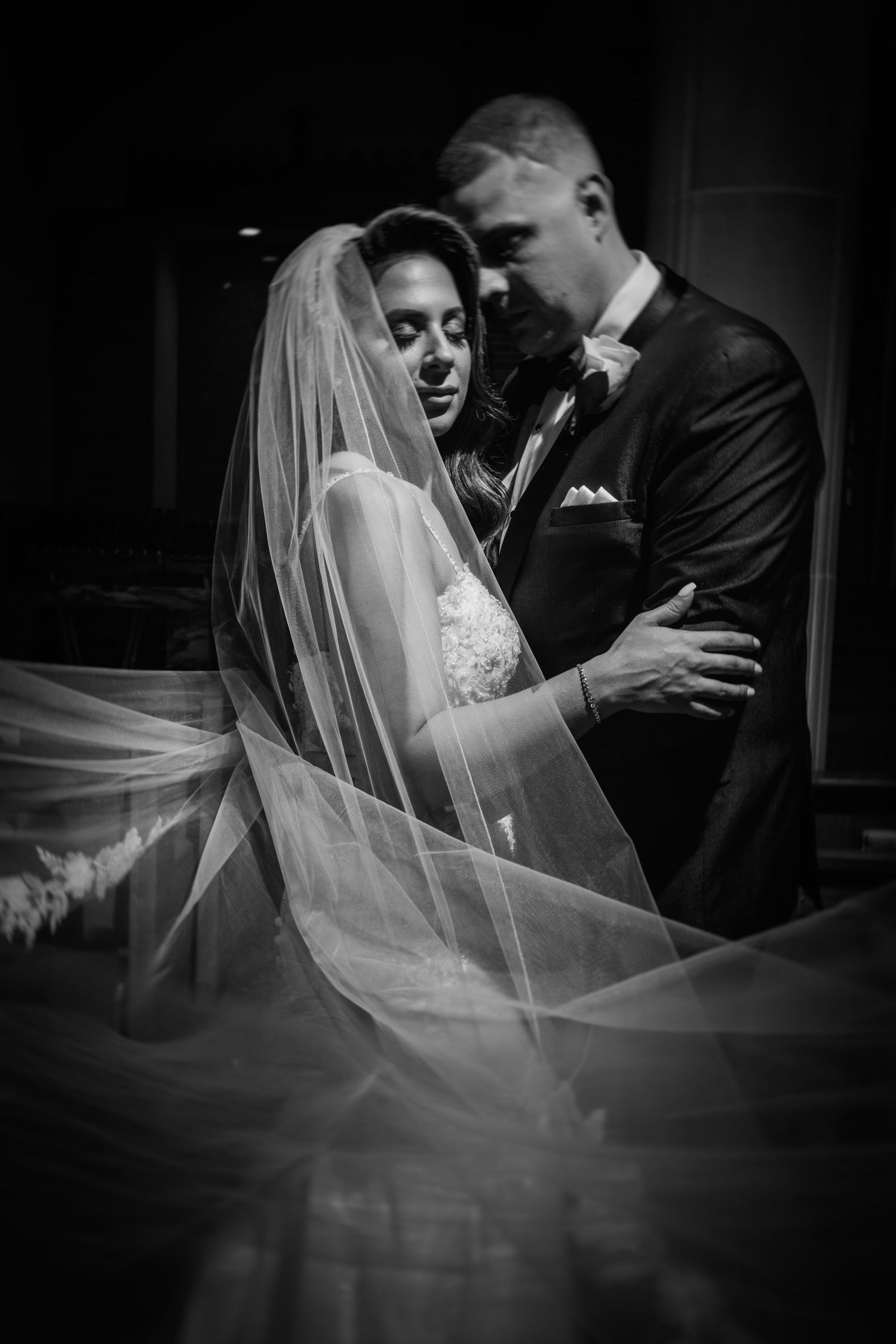 A romantic black and white wedding photo of a couple embracing with the bride wearing a flowing veil.