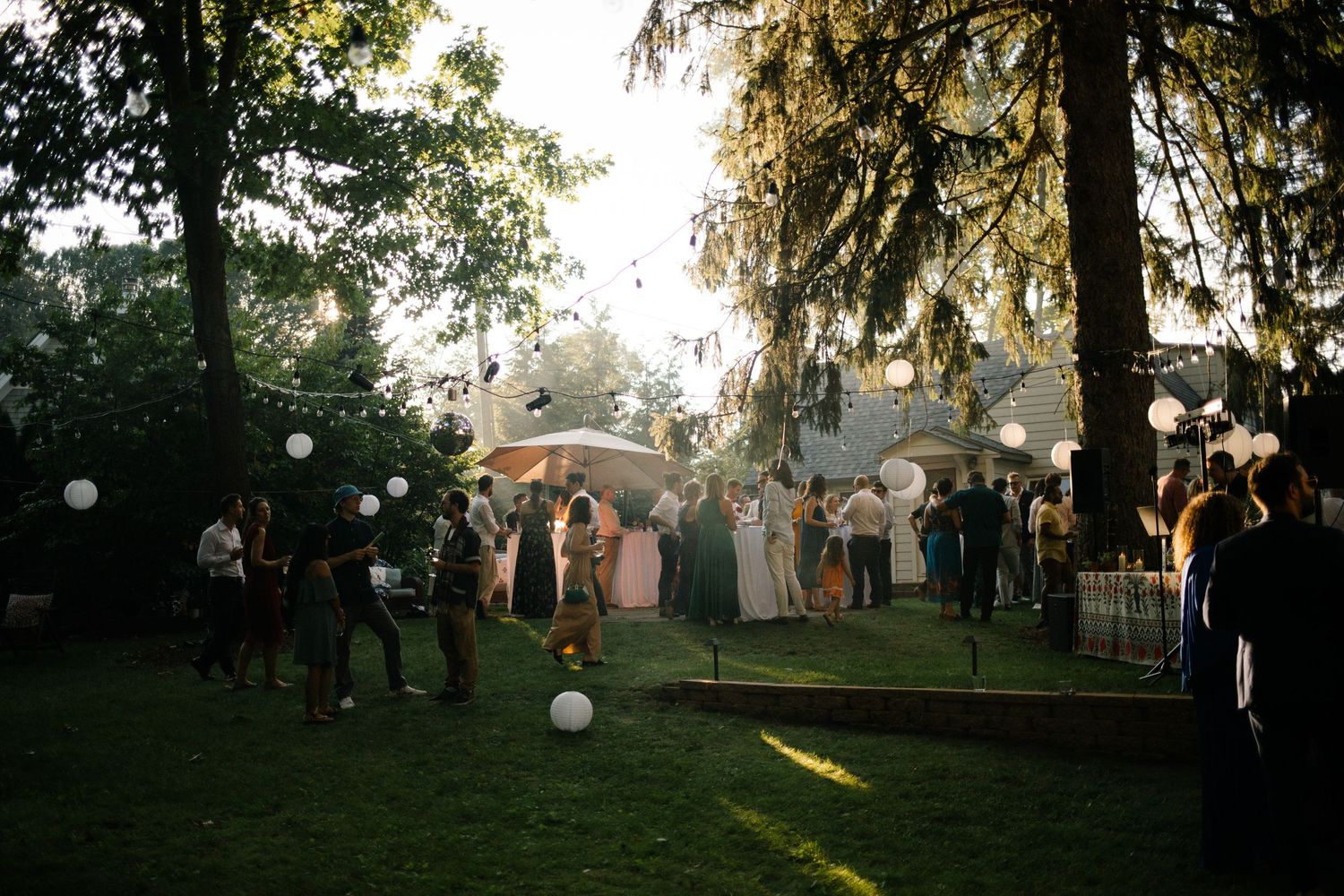 Outdoor wedding reception at dusk with guests mingling on a lawn with globe lights.