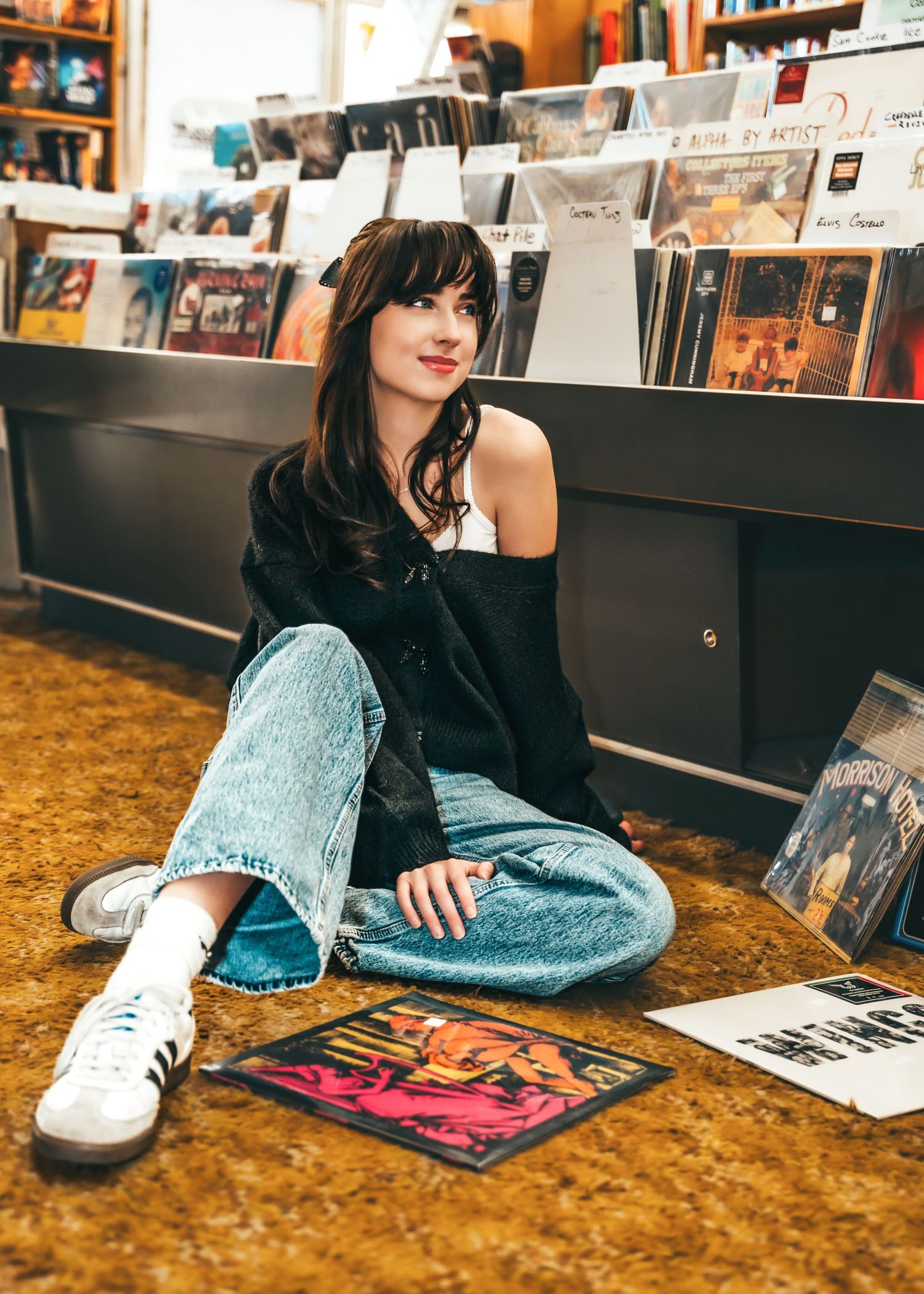 A figure stands between wooden bookshelves in a cozy library with warm lighting and hardwood floors.