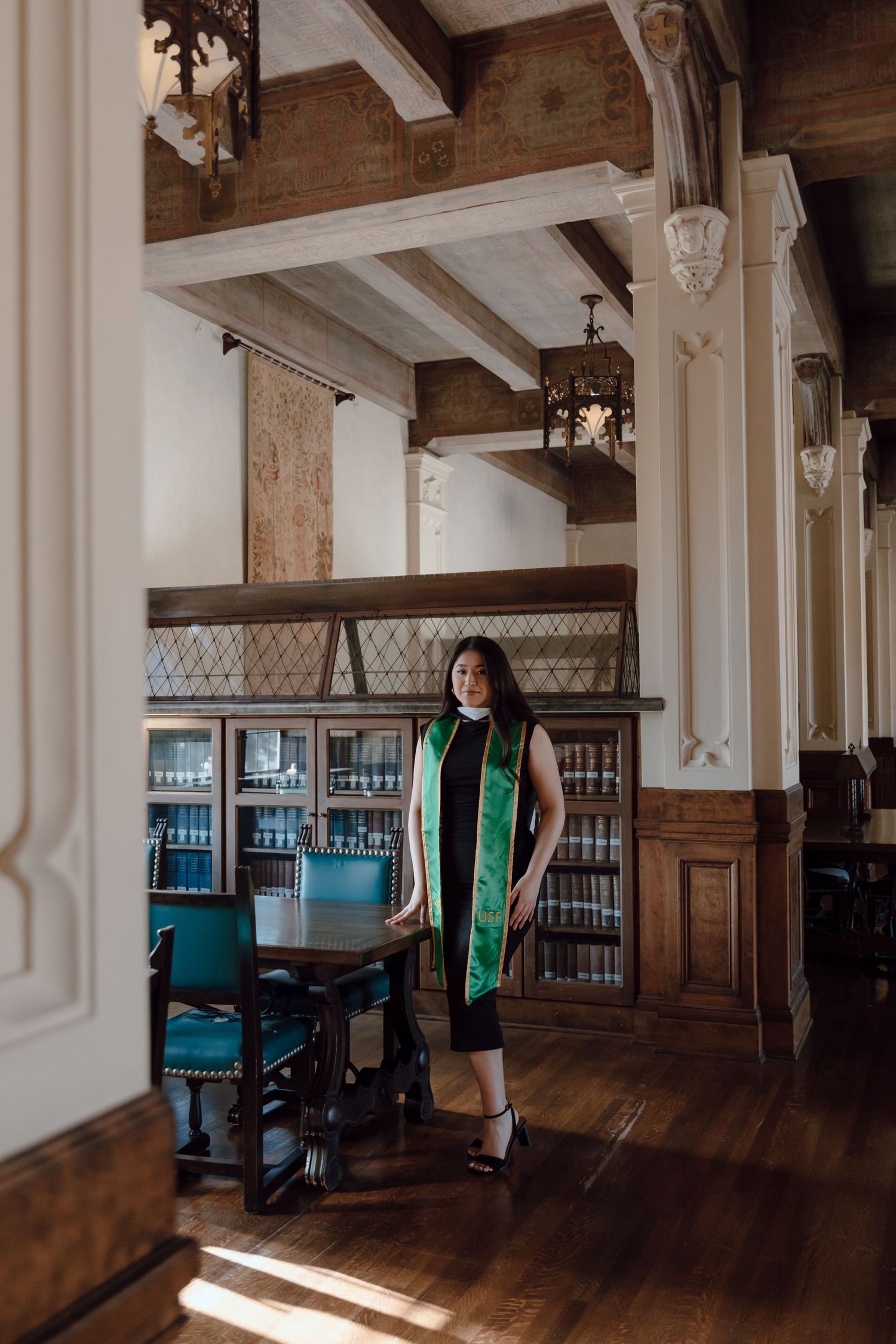 Graduate stands in the library at the University of San Francisco. Bookshelves and wood surround her. By Bay Area Graduation Photographer.